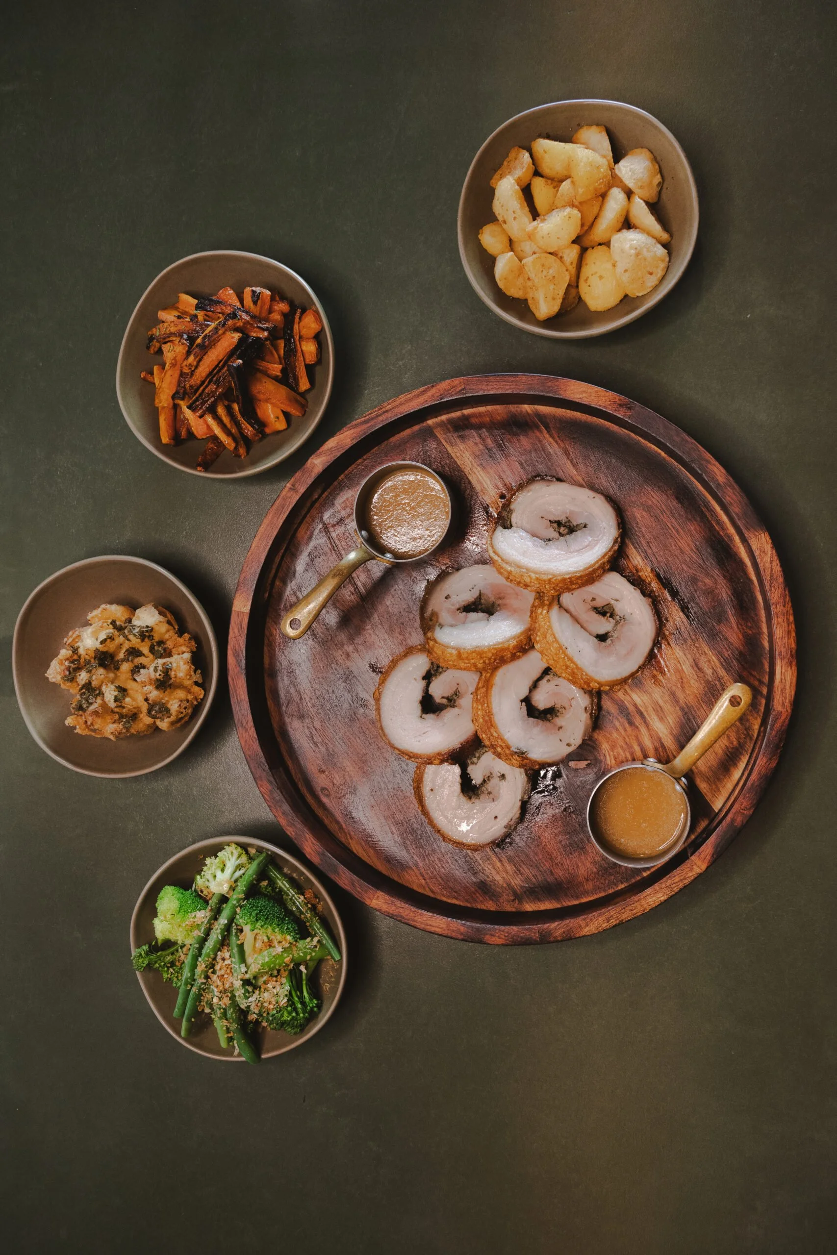 A table with a wooden tray of sliced rolled chicken with stuffing, small bowls of mashed potatoes, roasted carrots, broccoli, and green beans.