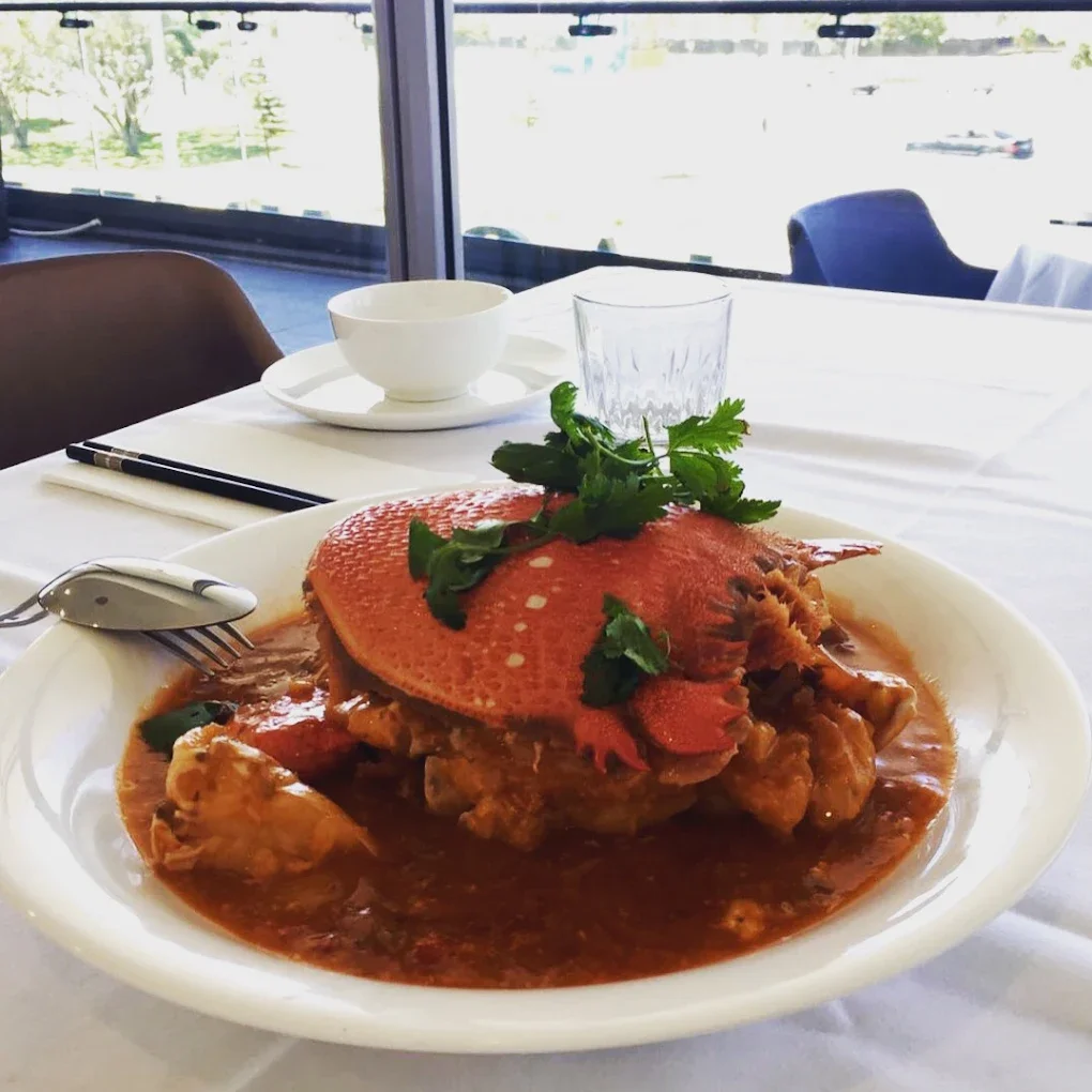 A plate of Chinese-style braised meat with a large red flower-shaped garnish and fresh cilantro on top, served in a sauce. The table is set with chopsticks, a spoon, a white bowl, a clear glass, and a cup. The background shows large windows with a view of trees and a parking lot.