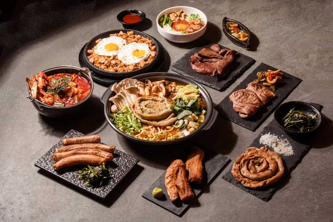 Assorted Korean dishes including grilled meats, soups, kimchi, rice, and side dishes arranged on black and gray serving plates and bowls on a dark table.