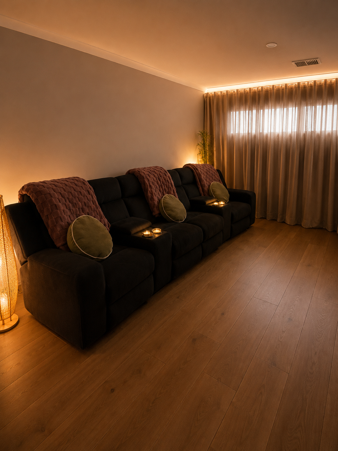 Living room with a black couch decorated with throw pillows and blankets, warm lighting, wooden floor, and curtains covering a window.