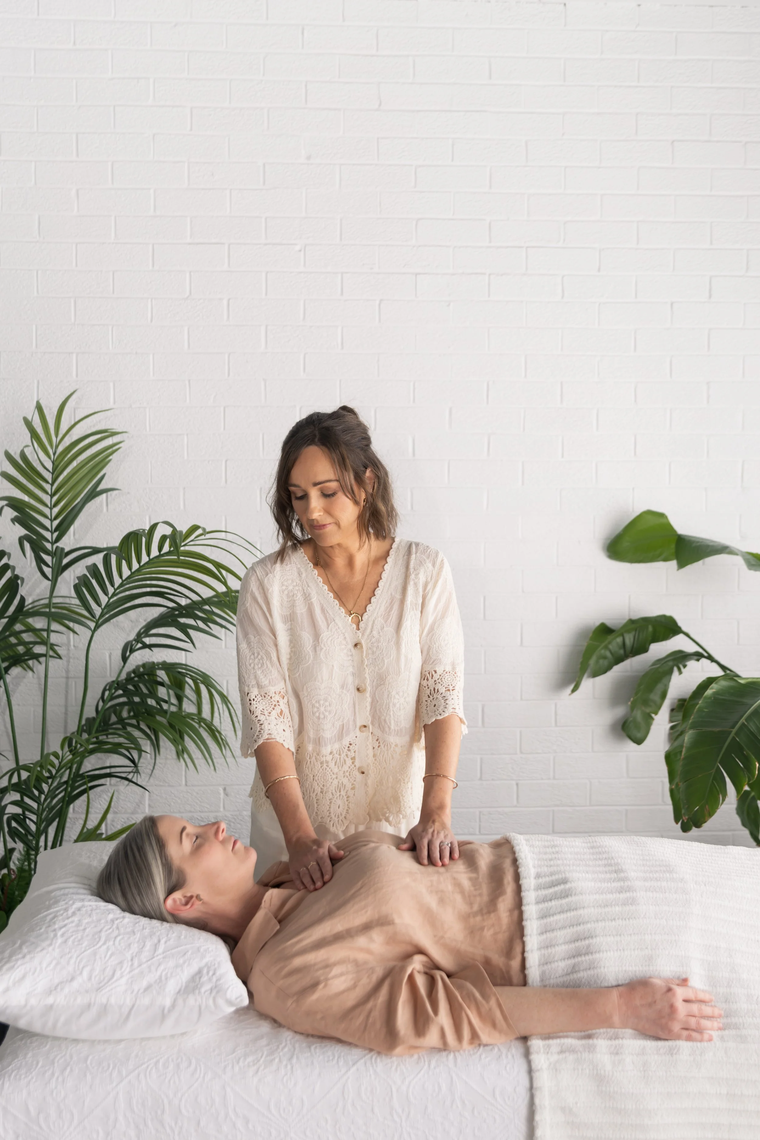A woman lying on a bed receiving a massage from another woman in a room with white brick wall and green plants.