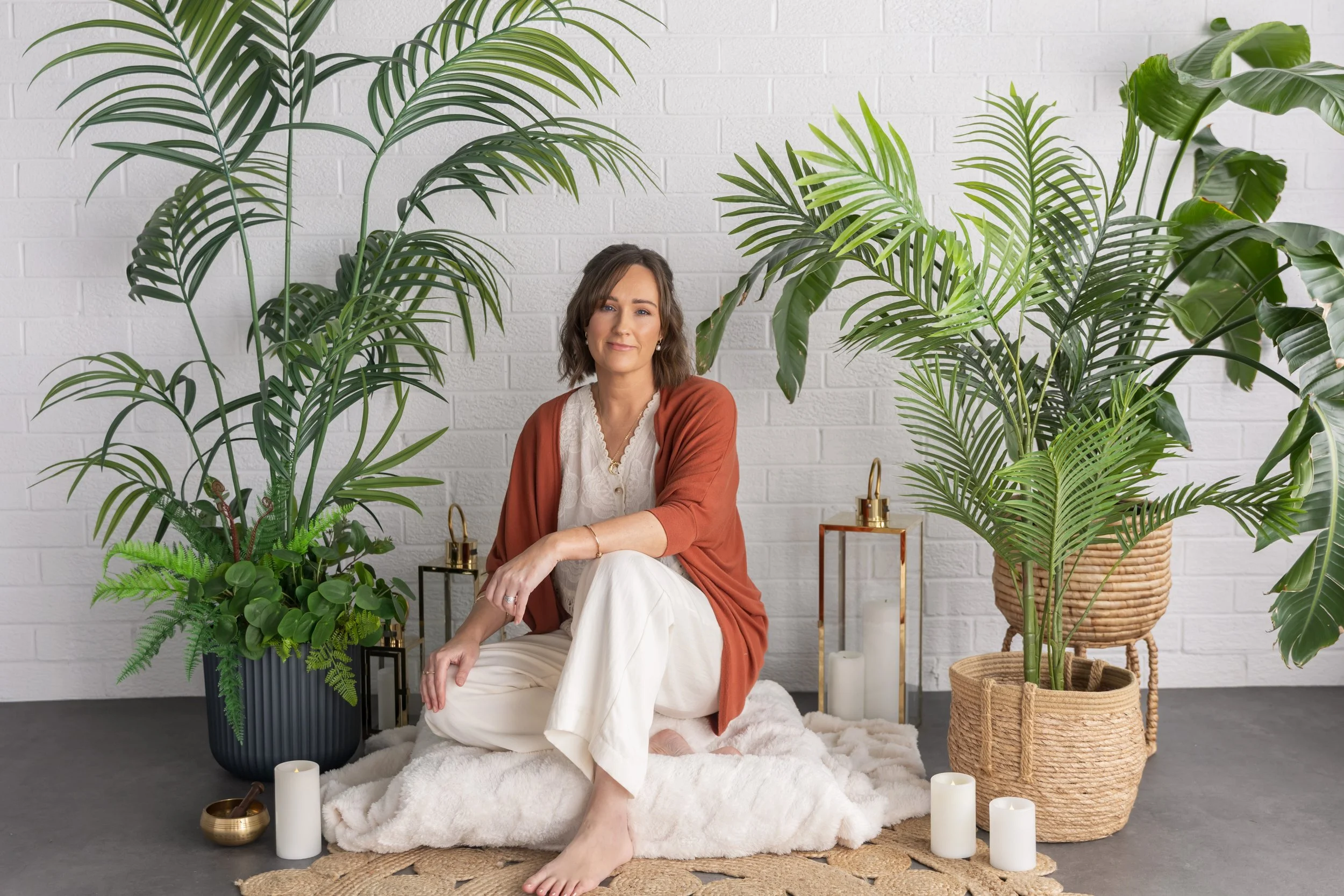 A woman sitting on a cozy white blanket surrounded by various large potted green houseplants, candles, and decorative objects in a bright room with a white brick wall.