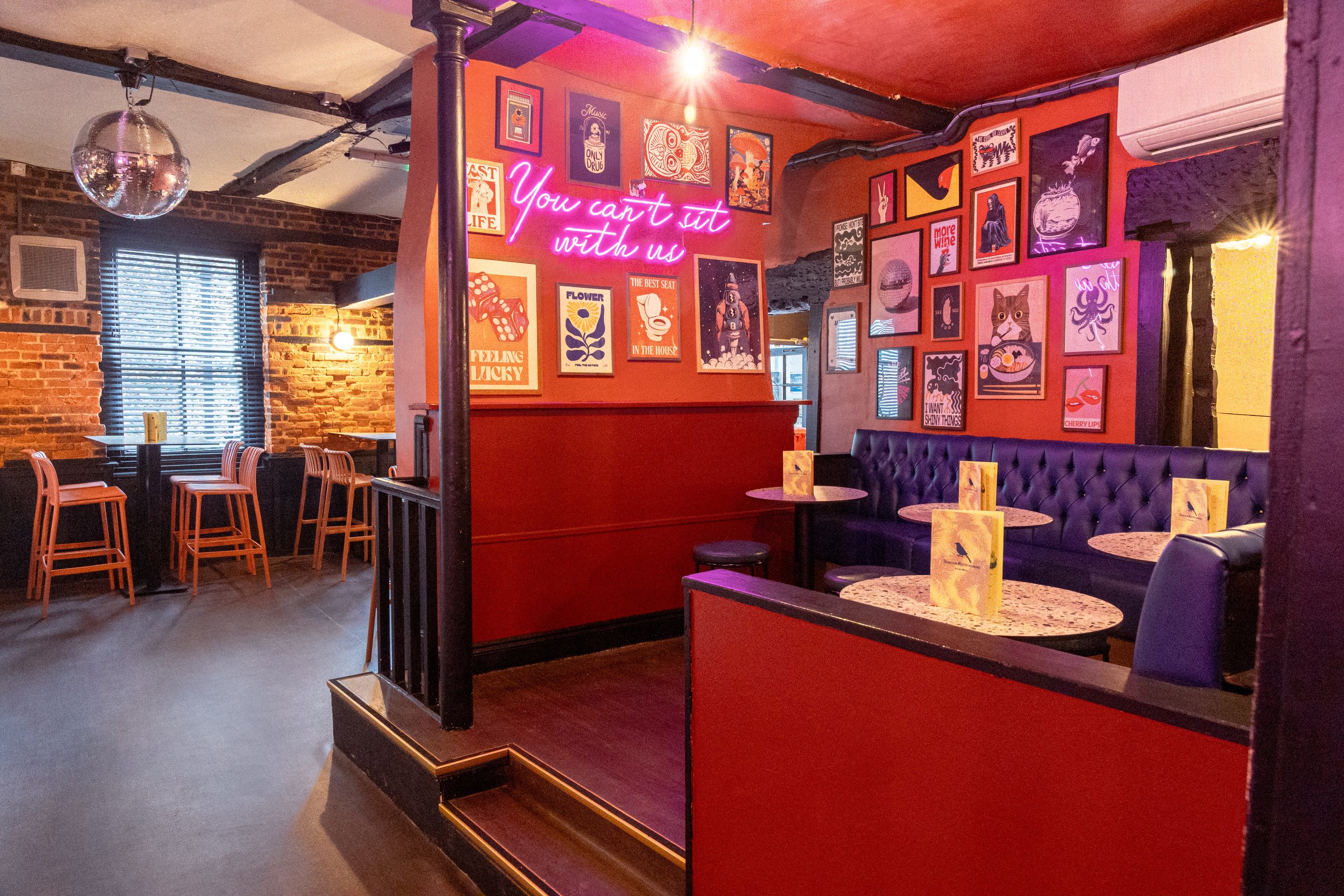 Interior of a cafe or restaurant with a neon sign that says "You can't sit with us." The space has exposed brick walls, framed artwork, a disco ball, and a cozy seating area with purple tufted banquettes and small round tables. There are additional chairs along the windows, which have blinds. The decor features warm lighting and colorful art, creating a trendy, artistic atmosphere.