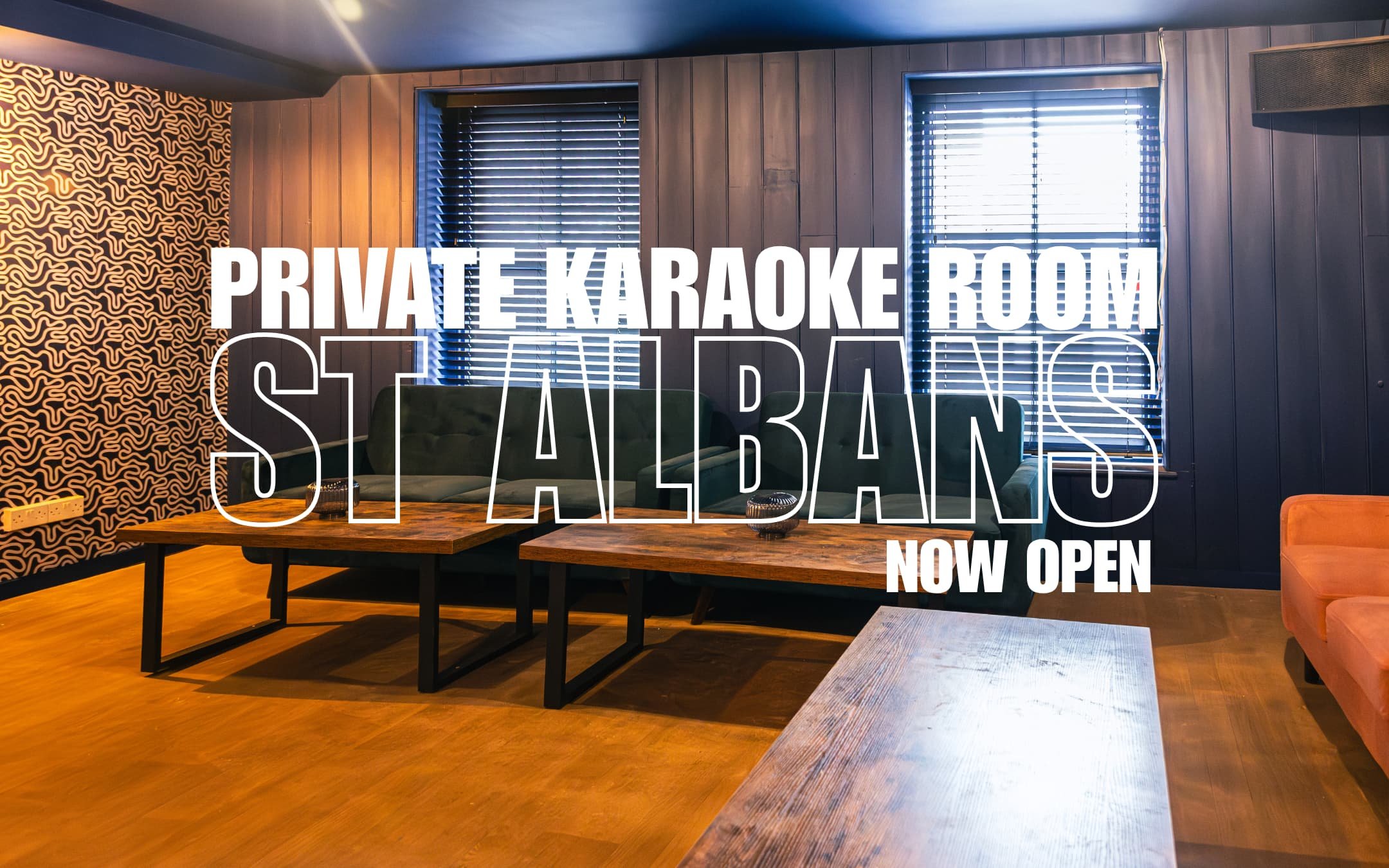 Interior of a private karaoke room with wooden floors, dark wood-paneled walls, and windows with blinds. There are green velvet sofas, a wooden table, and a framed sign that reads "Private Karaoke Room St Albans Now Open."