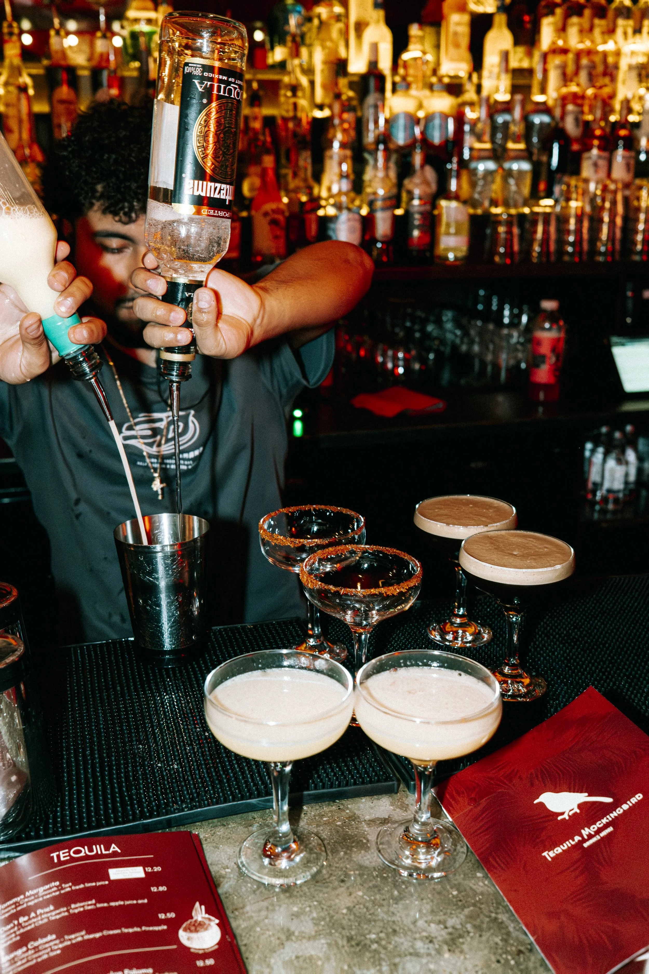 Four dark-colored cocktails with frothy tops in coupe glasses on a pink surface.