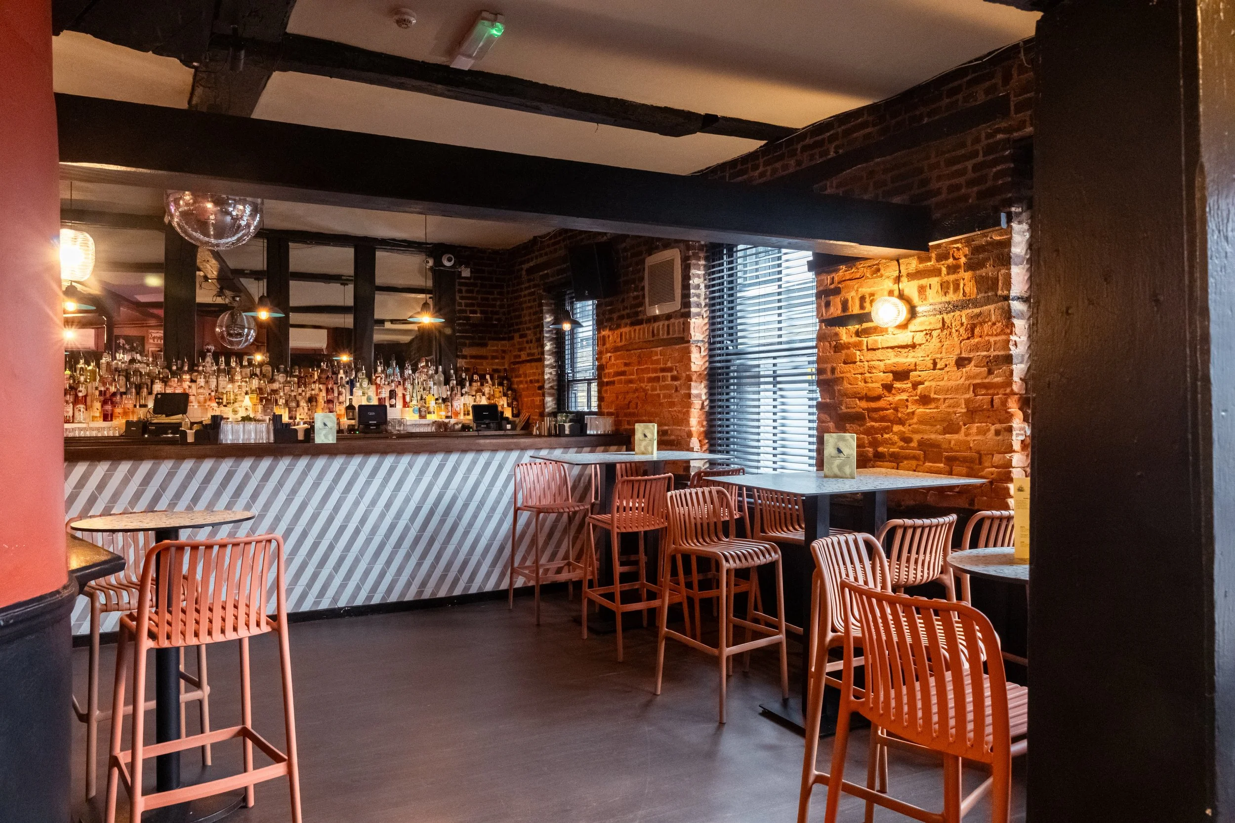 Interior of a modern bar with exposed brick walls, a curved bar counter filled with liquor bottles, and salmon-colored chairs around tables near large windows with blinds.