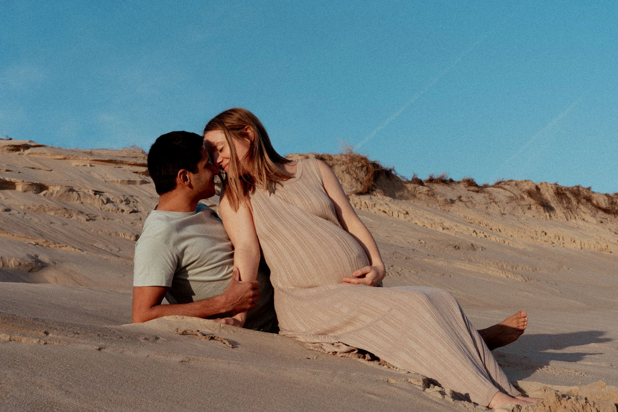 Un couple alité sur des dunes de sable dans un désert, elle est enceinte, ils se regardent tendrement sous un ciel bleu.