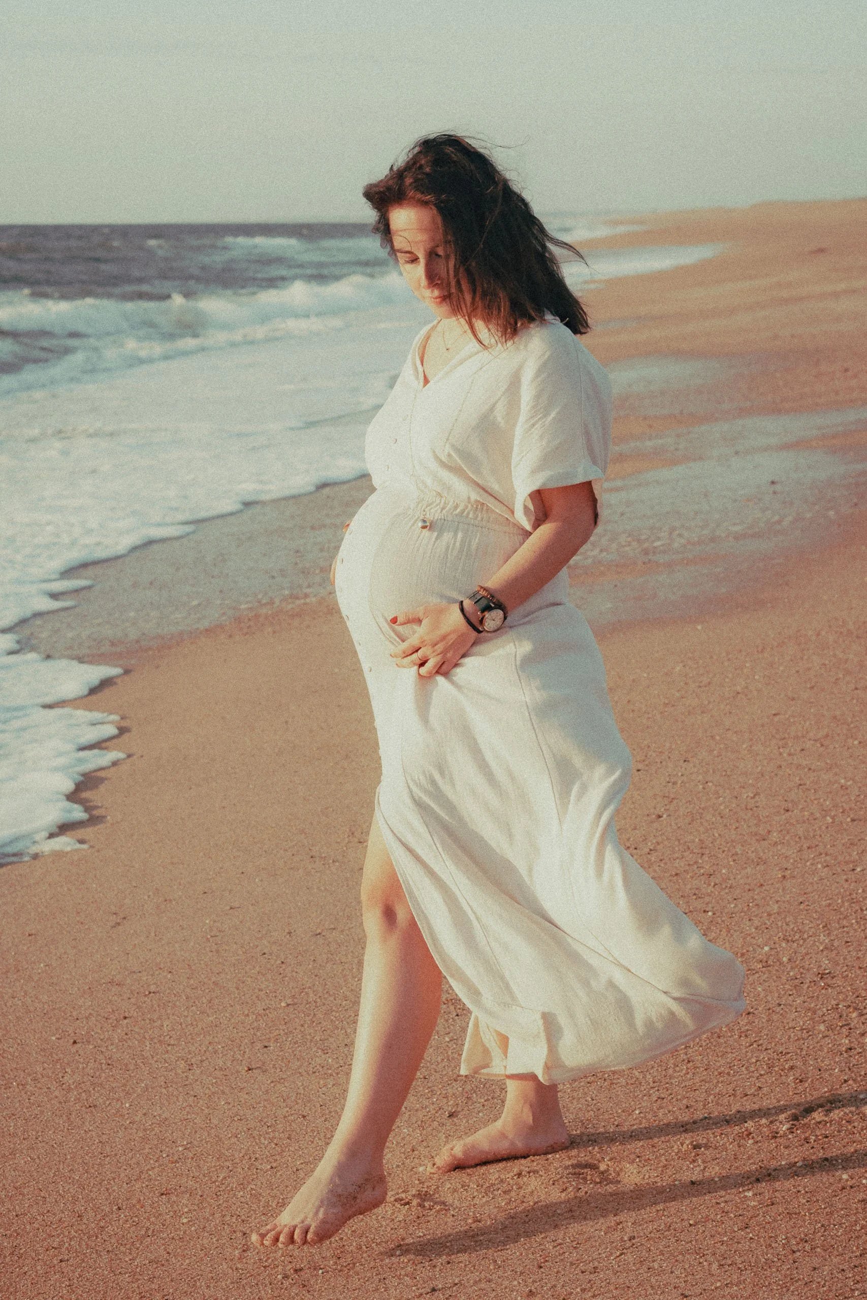 Femme enceinte marchant pieds nus sur la plage, portant une robe blanche