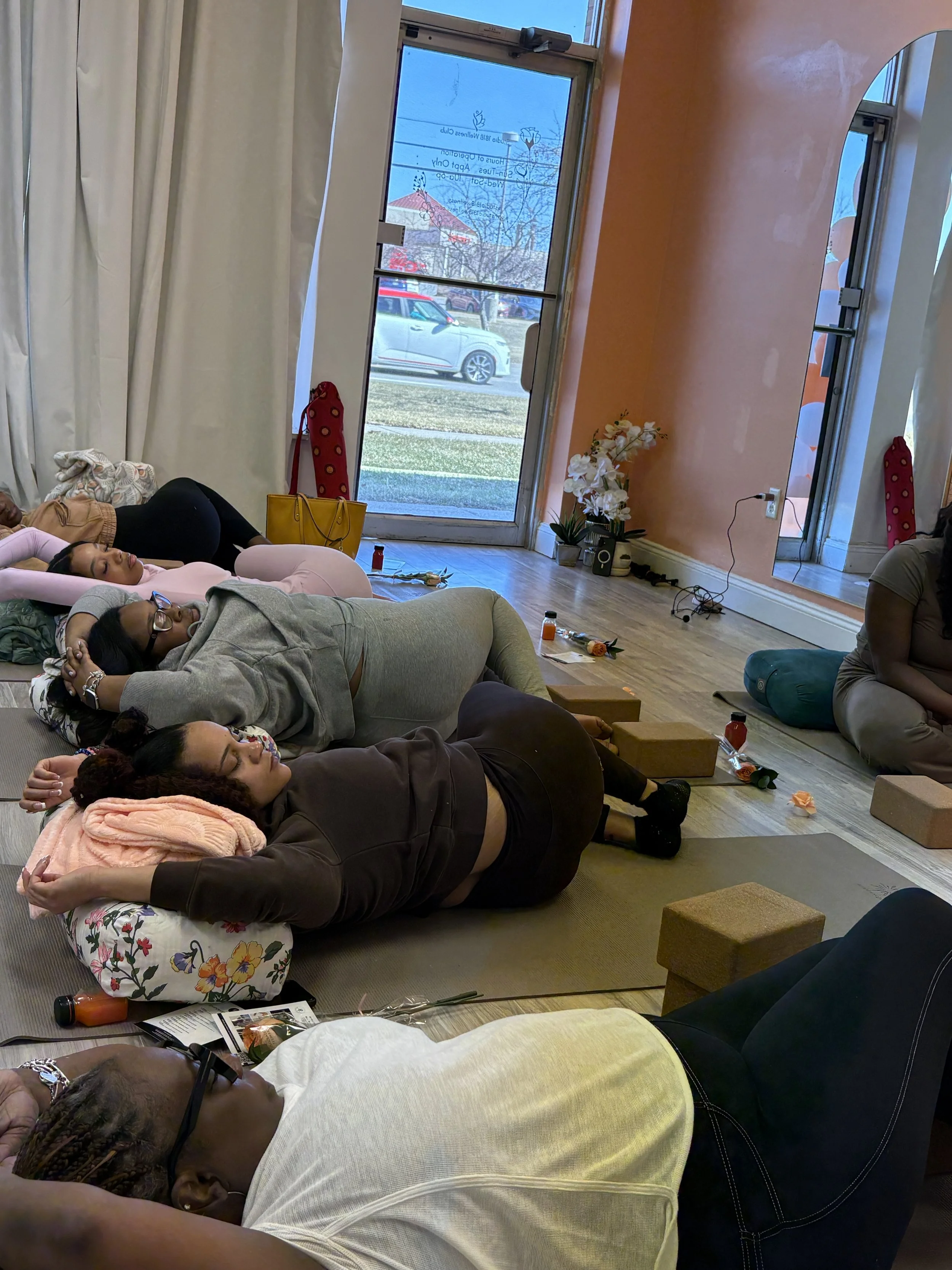 People lying down and relaxing on mats during a meditation or relaxation session in a bright room with orange and white walls, large windows, and wood flooring.