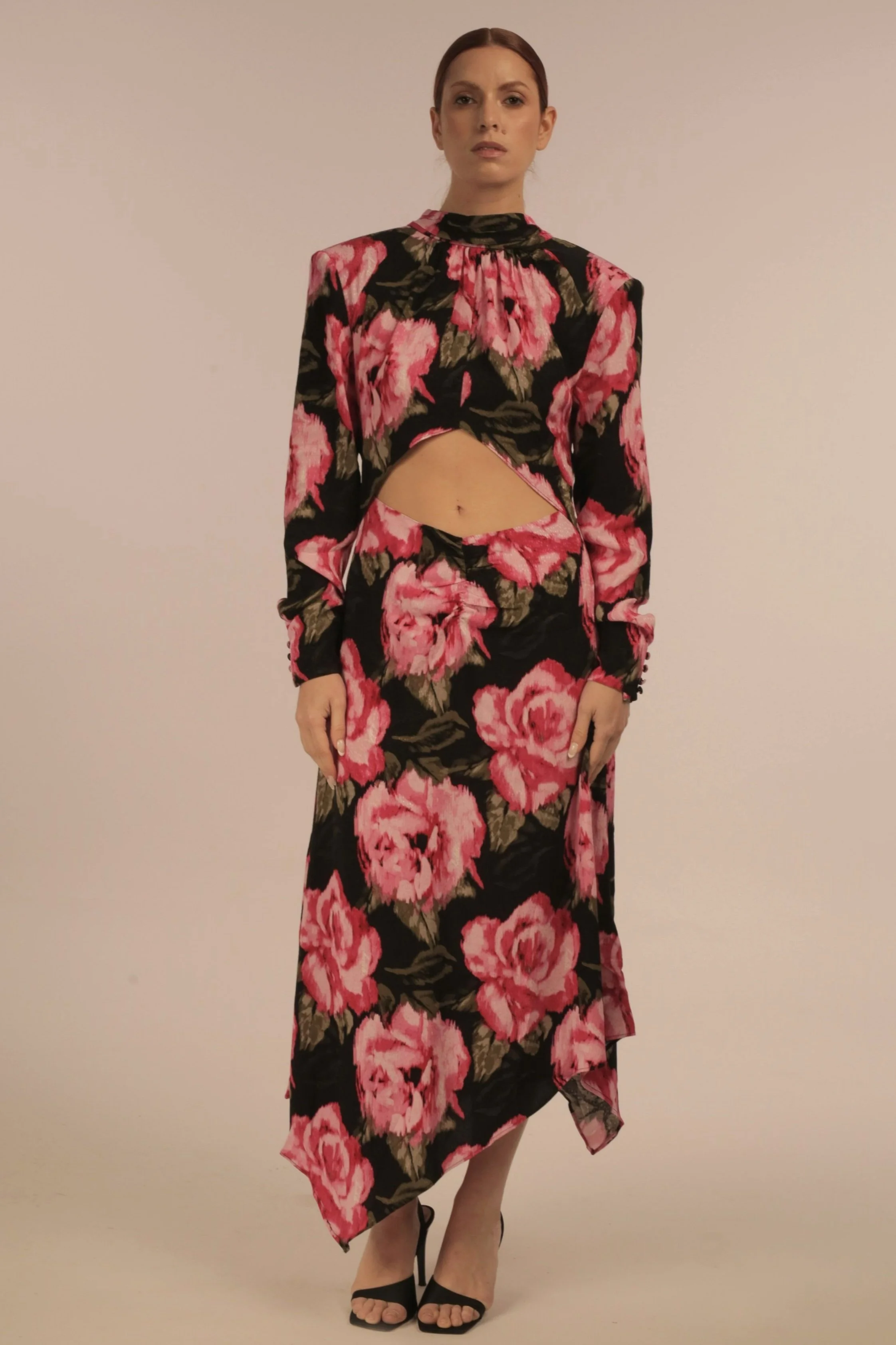 A woman standing against a plain background, wearing a matching floral two-piece outfit with pink roses on black fabric, black high heels, and her hair pulled back.