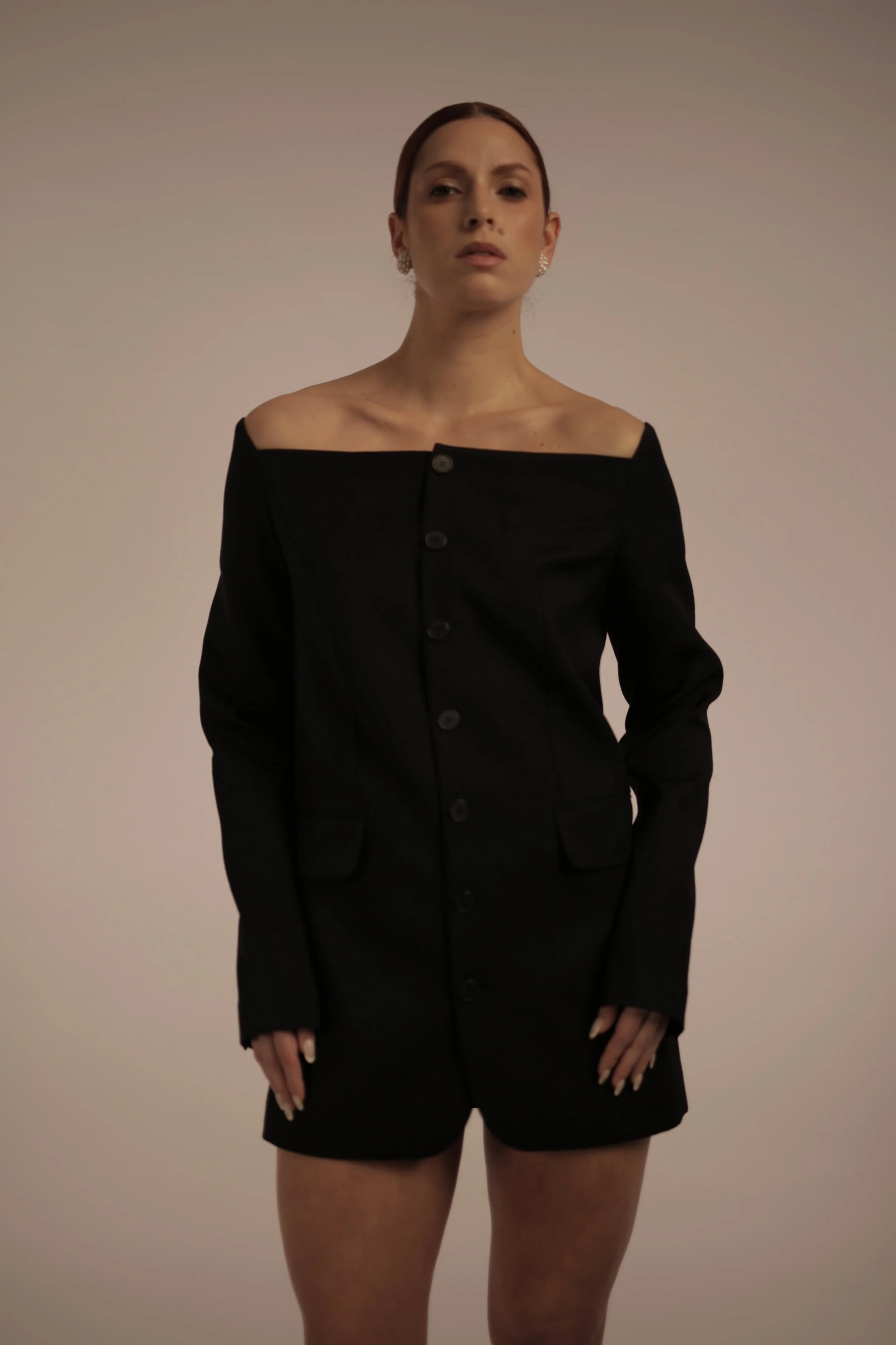 Woman with red hair wearing a black off-the-shoulder blazer and shorts, standing against a plain background.