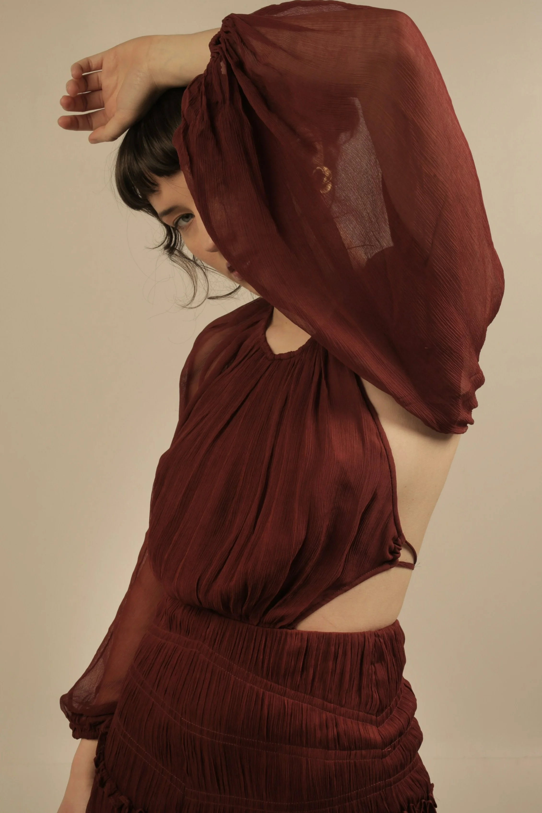 Woman wearing a sheer, rust-colored dress with cut-out side, posing with her left arm raised above her head.