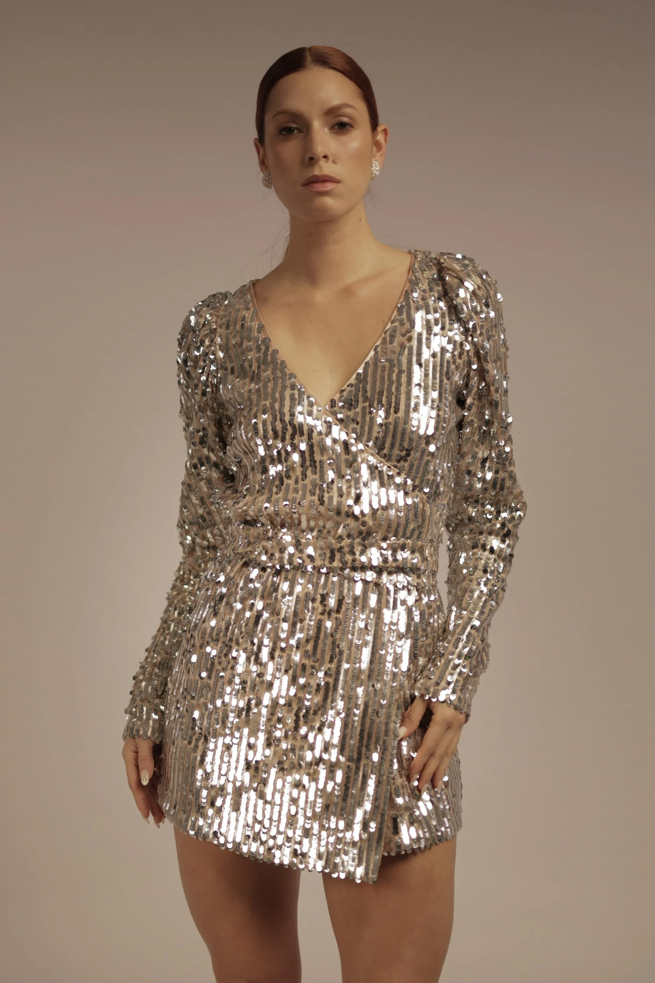 A woman with brown hair in a low bun wearing a gold sequined dress with long sleeves and a wrap-style V-neck.