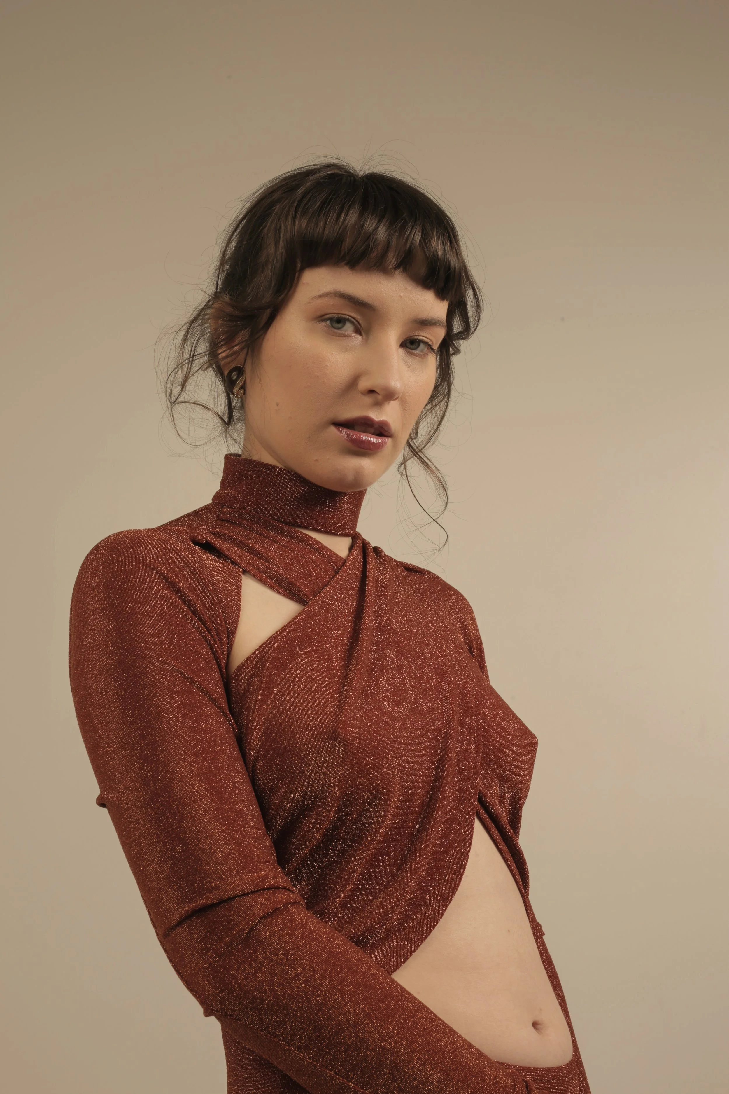 A woman with short brown hair and bangs wearing a rust-colored, shimmery dress with cutouts, standing against a plain beige background.
