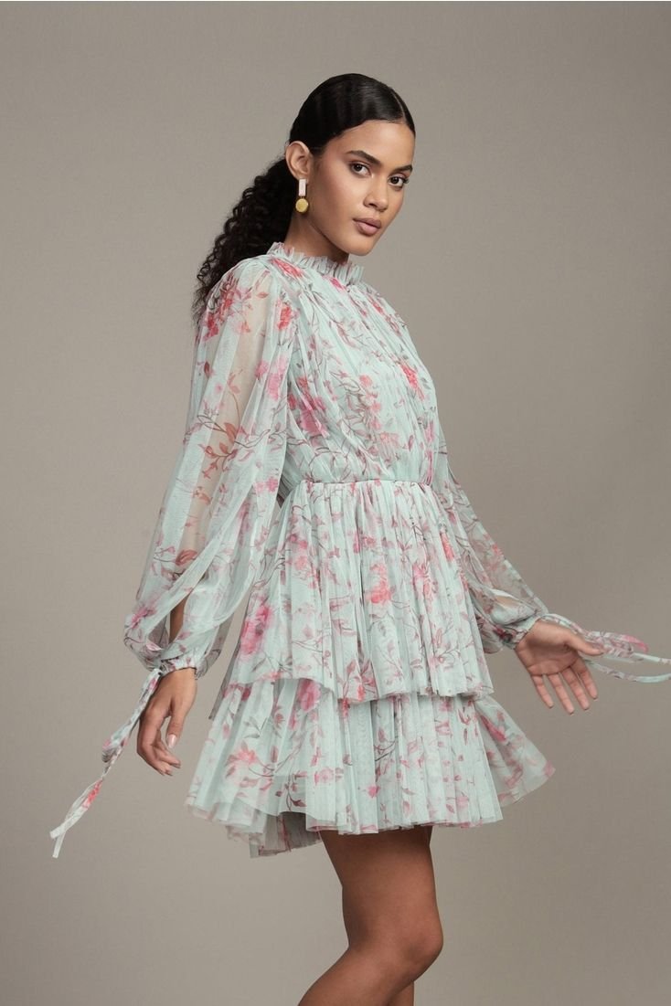 A woman wearing a flowy, floral dress with long sleeves and a tiered skirt, standing against a plain background.