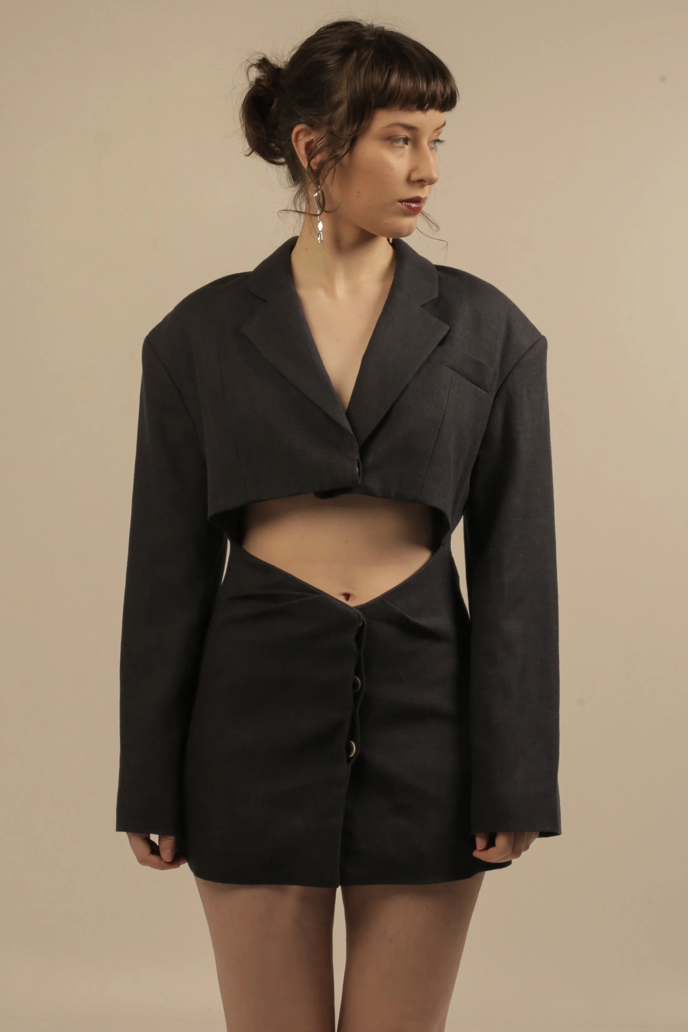 A young woman with brown hair in a messy bun, wearing a black cropped suit jacket and a matching high-waisted skirt with buttons, standing against a plain light-colored wall.