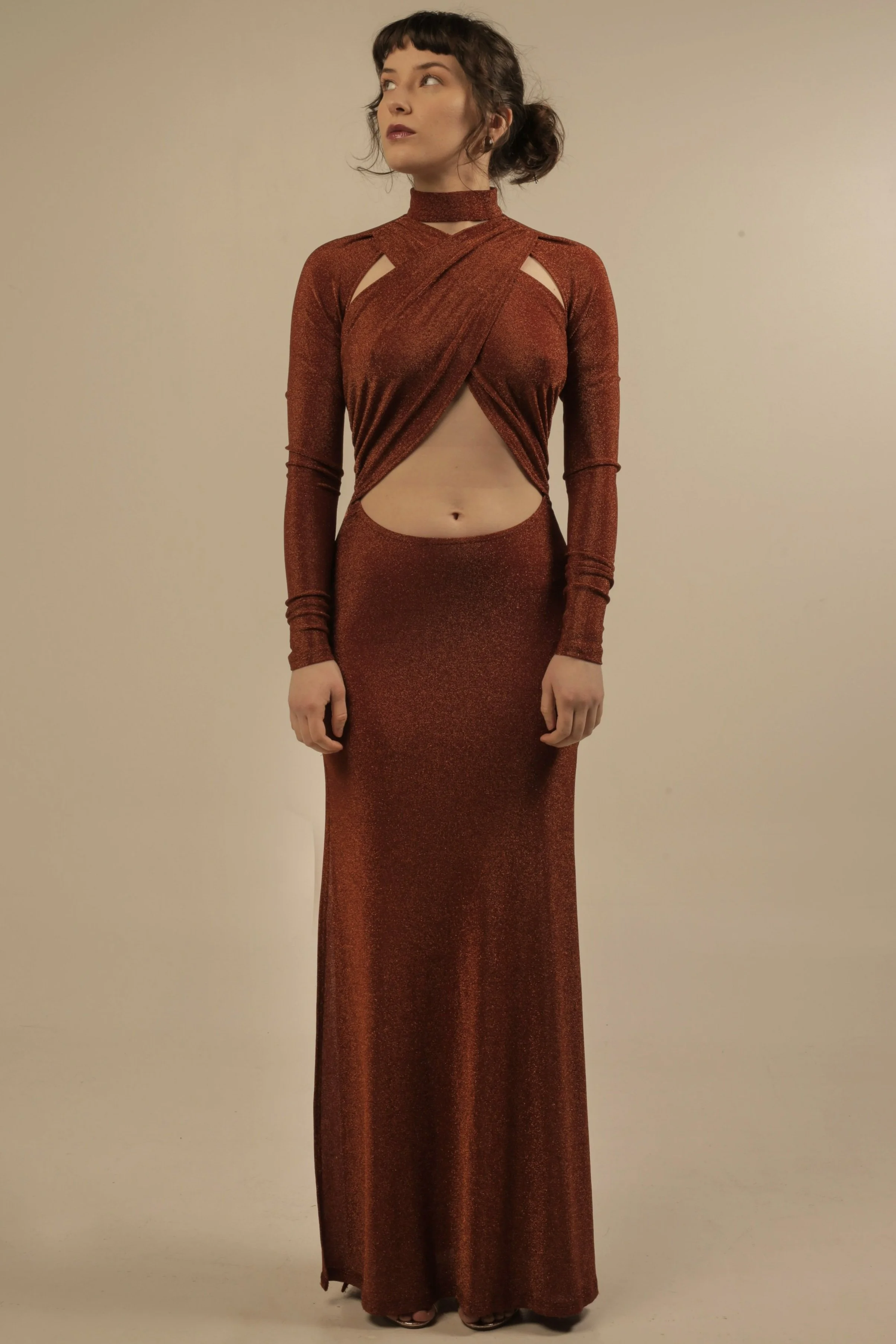 A woman wearing a long, shiny, rust-colored dress with cut-out details on the shoulders and midriff, standing against a plain background.
