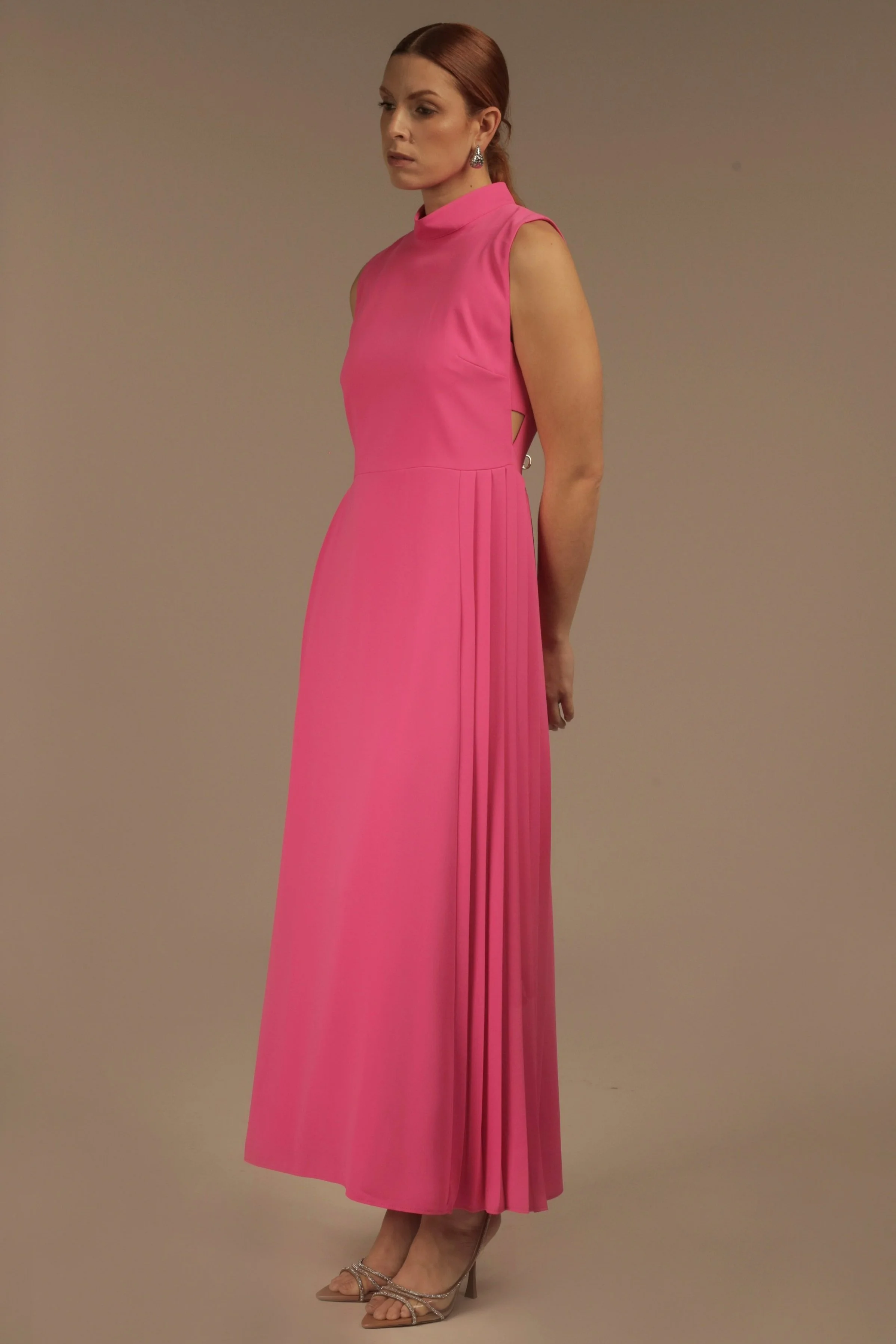 A woman wearing a long, sleeveless, pink evening gown with a high neckline, standing against a plain background. She has reddish-brown hair styled back and is wearing earrings and high heels.