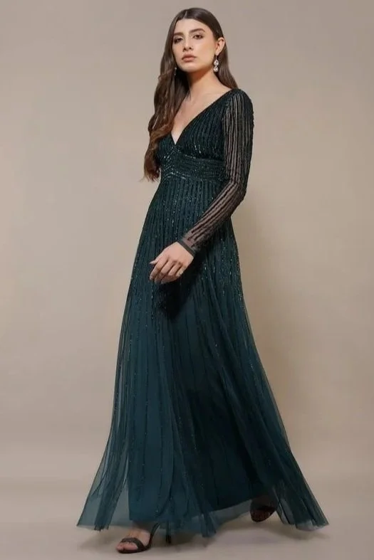 A young woman with long dark hair wearing a dark green, long-sleeved, pleated gown with a deep V neckline and earrings, posing against a plain neutral background.