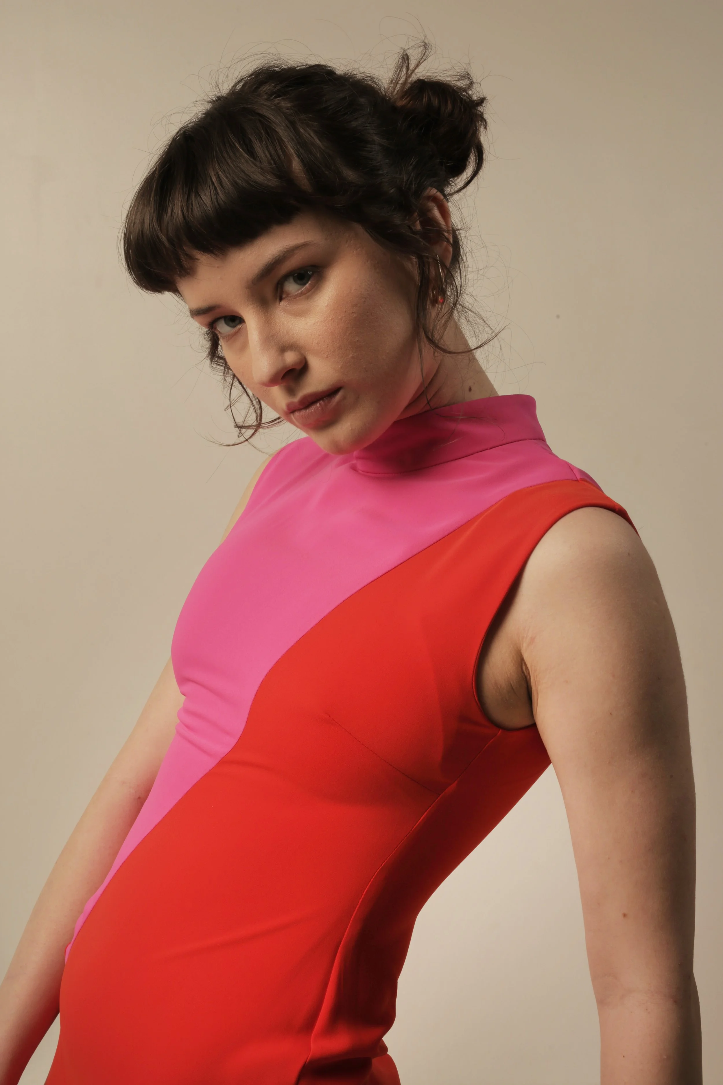 A young woman with short dark hair and bangs, wearing a sleeveless, high-neck dress with a color-block design in pink, red, and orange hues, posing against a plain neutral background.
