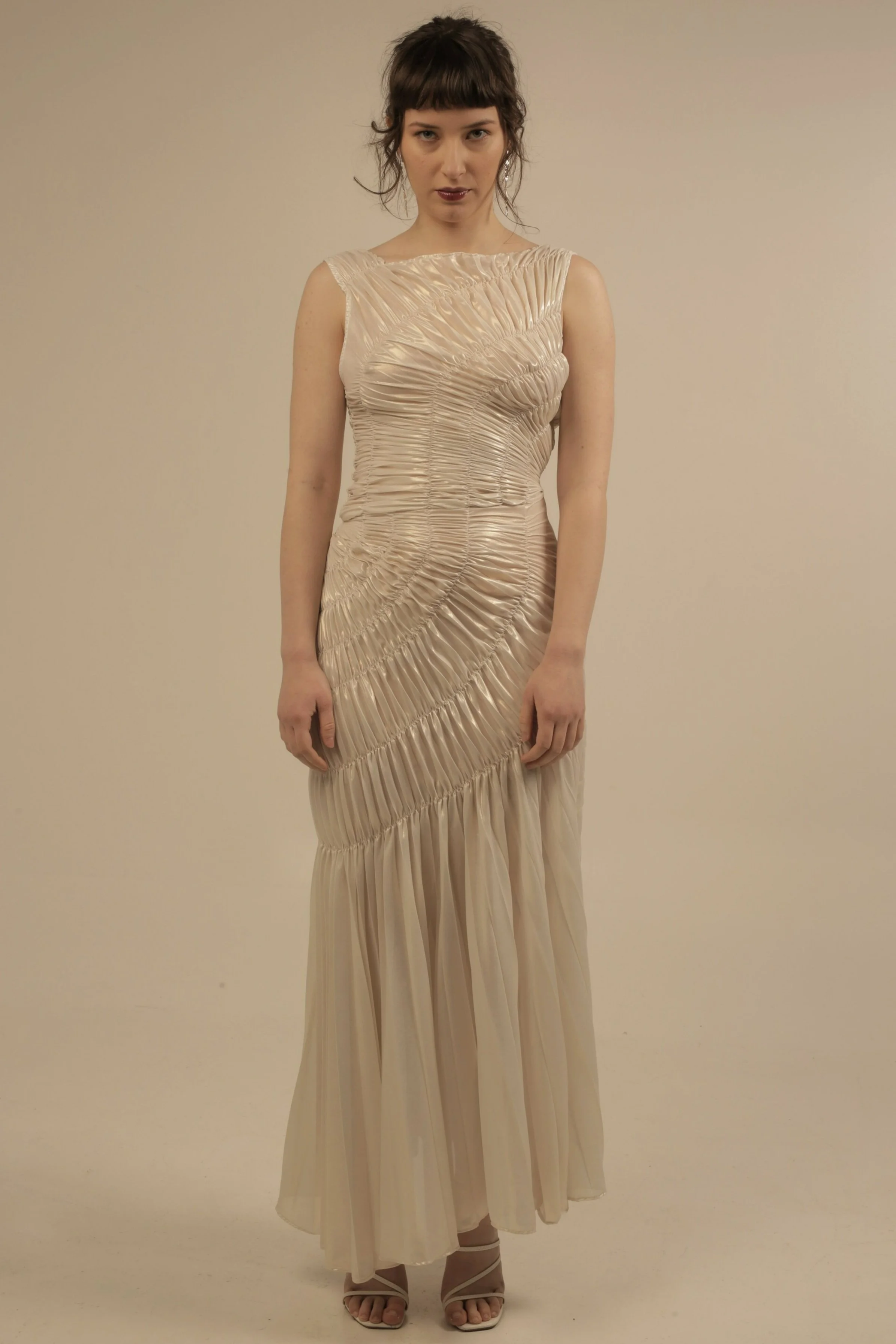 A woman with dark hair styled in loose waves and bangs, wearing a long, beige, pleated dress with a fitted bodice, standing against a plain background.