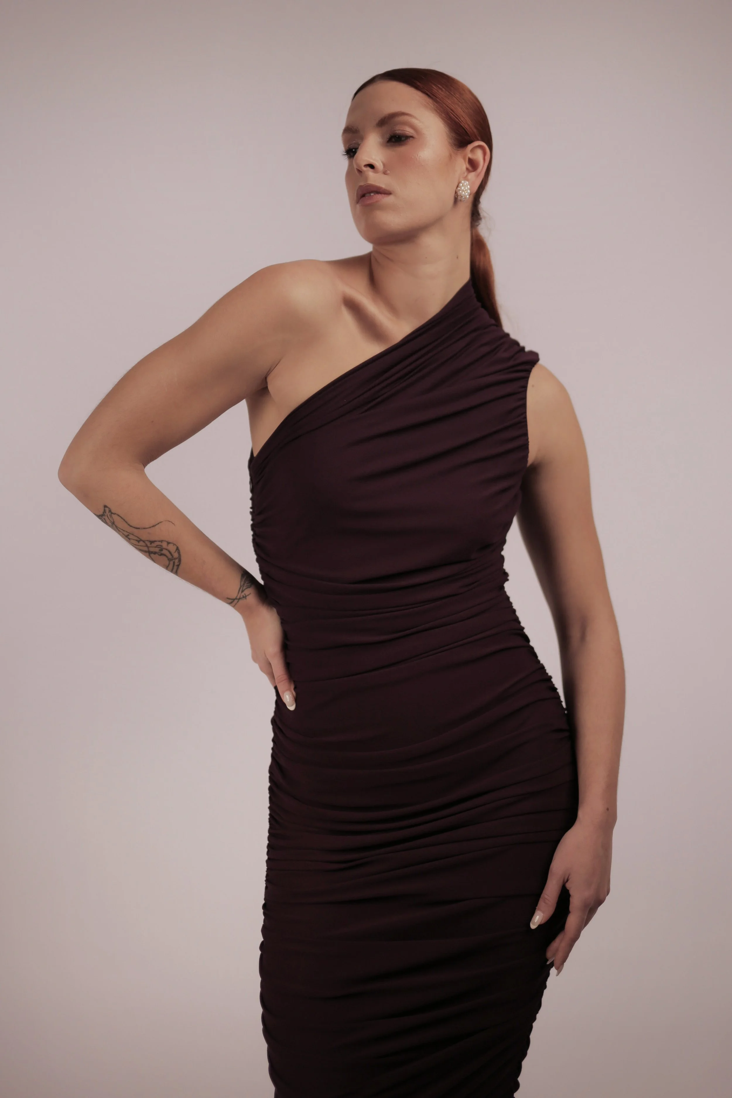 A woman with red hair in a ponytail wearing a one-shoulder dark purple ruched dress, earrings, and a tattoo on her left arm, standing against a plain light background.