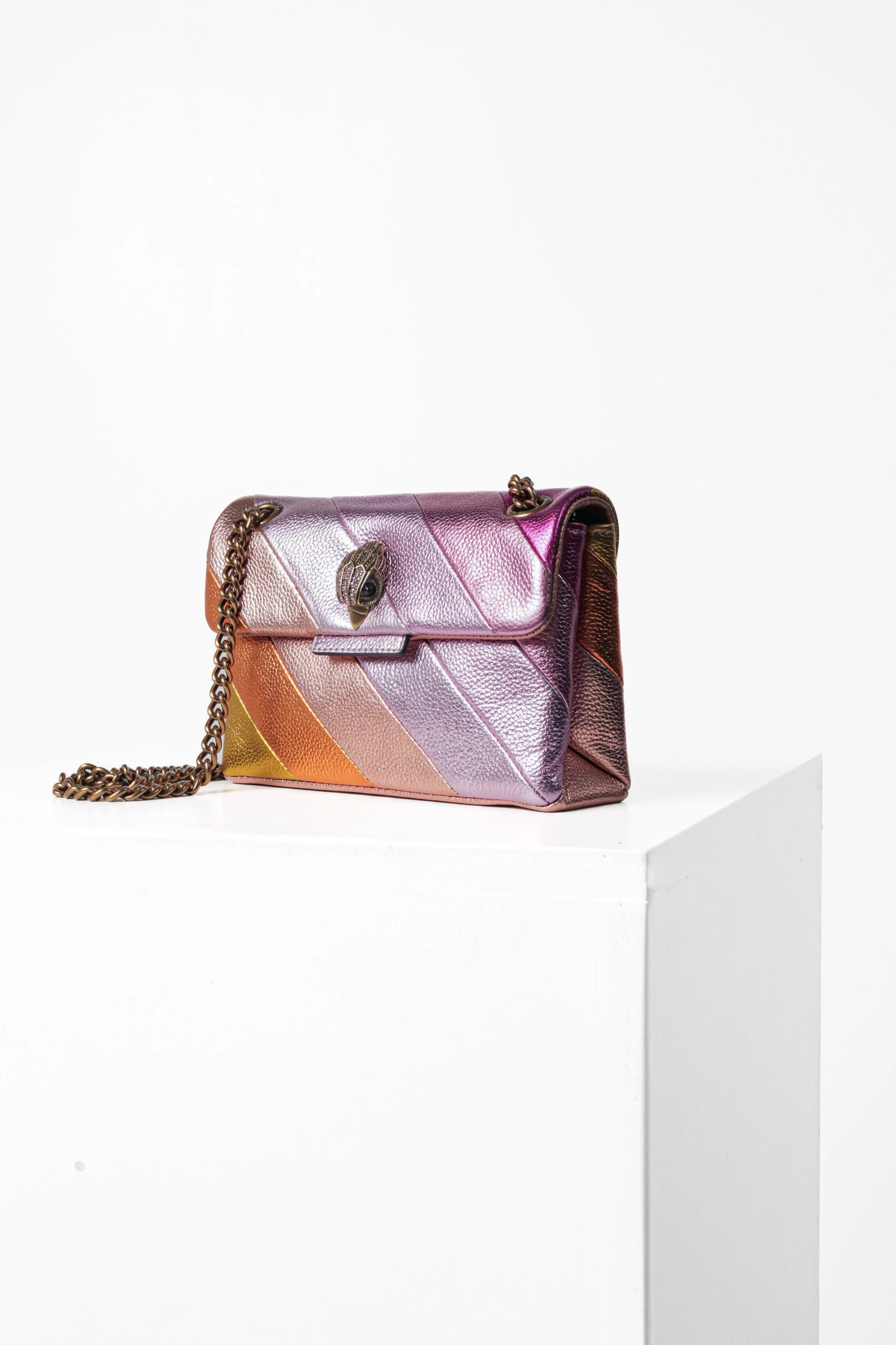 Iridiscent handbag with a chain strap and a eye-shaped clasp, displayed on a white pedestal against a white background.