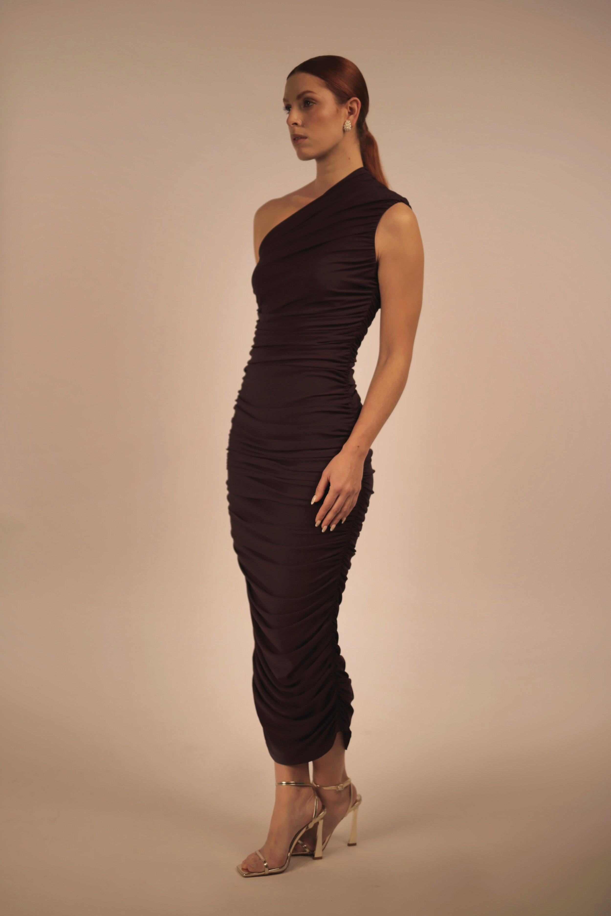 A woman with long, dark hair styled in a low ponytail, wearing a one-shoulder, dark ruched dress and high heels, standing against a plain, light-colored background.