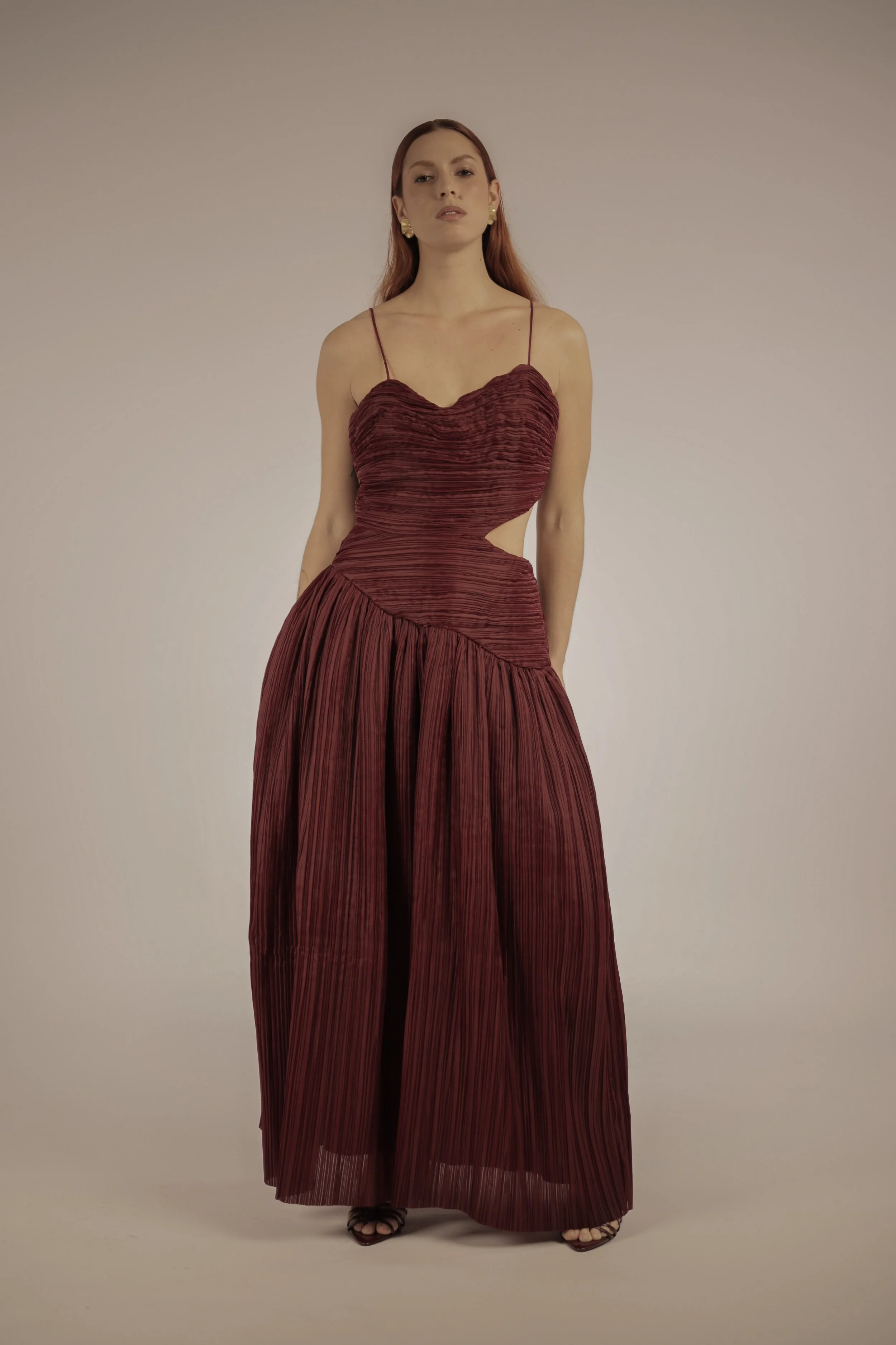 Woman wearing a red, pleated, cutout evening gown with spaghetti straps, standing against a plain background.