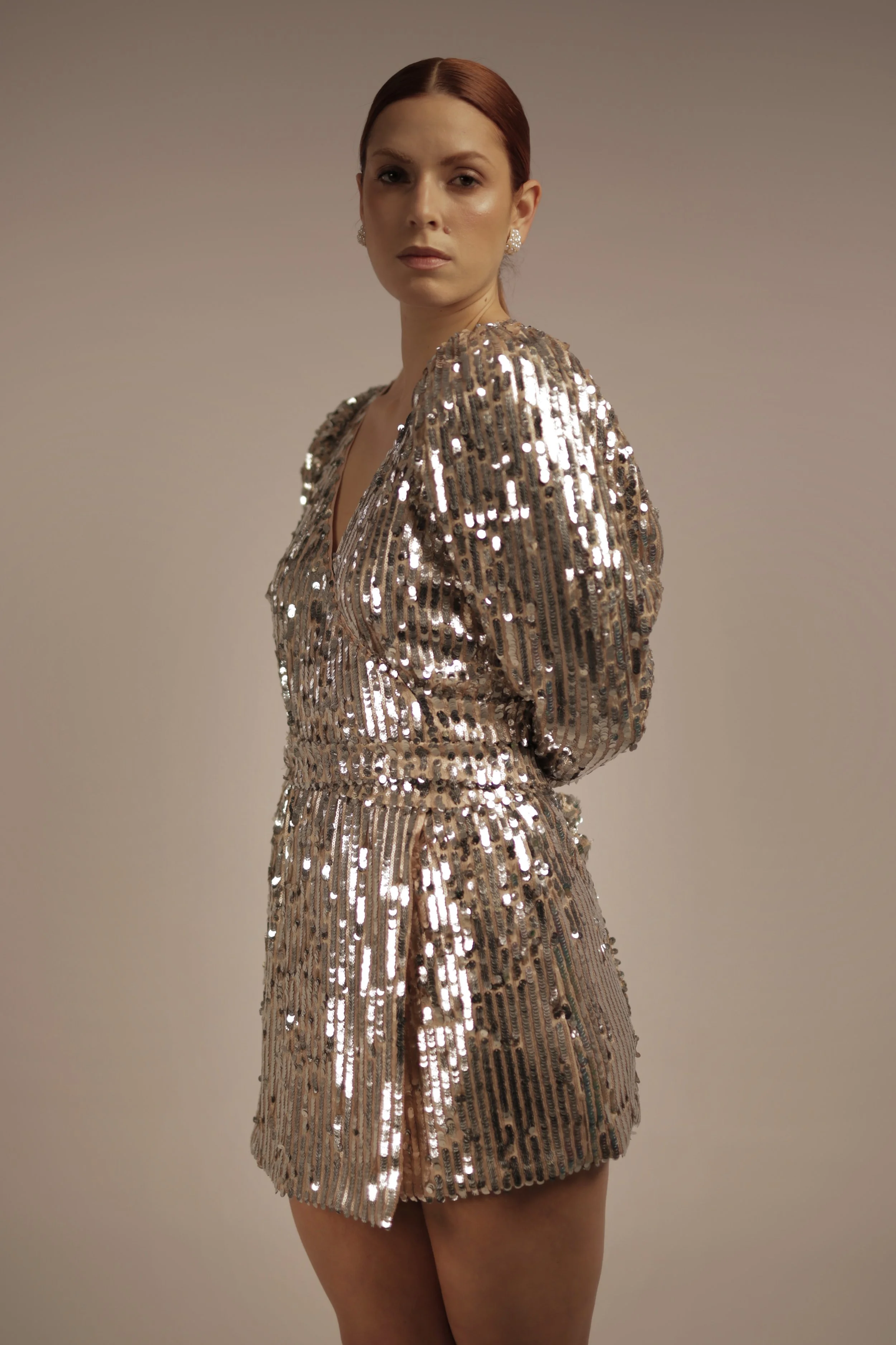 A woman with red hair wearing a shiny, sequined gold dress with long sleeves and a V-neck, standing against a plain background.