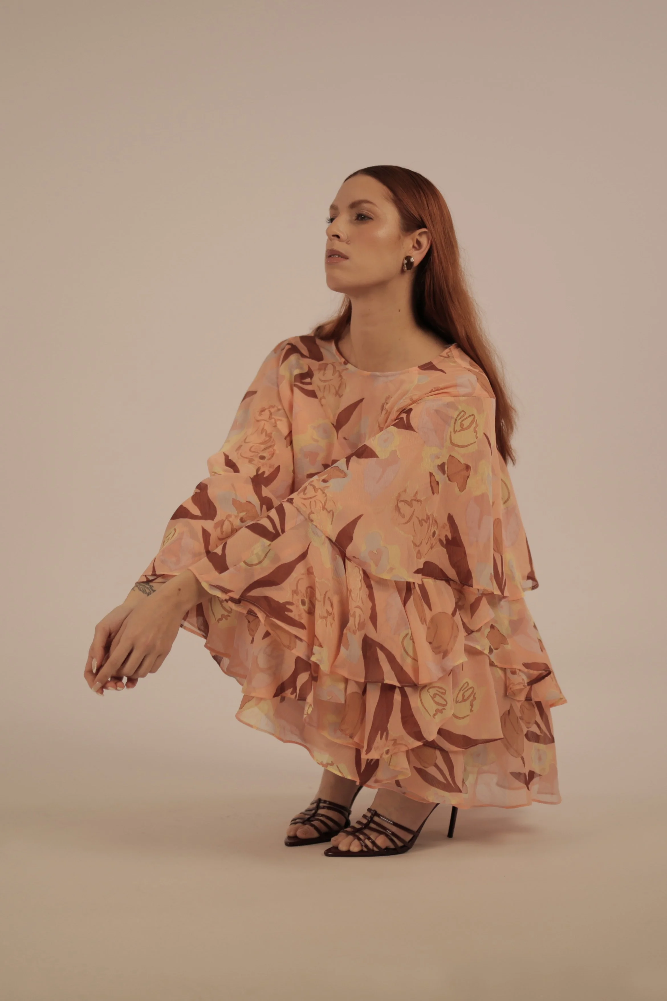 A woman with long red hair wearing a flowy, floral-patterned peach and brown dress and strappy high heels, squatting on the floor against a plain background.