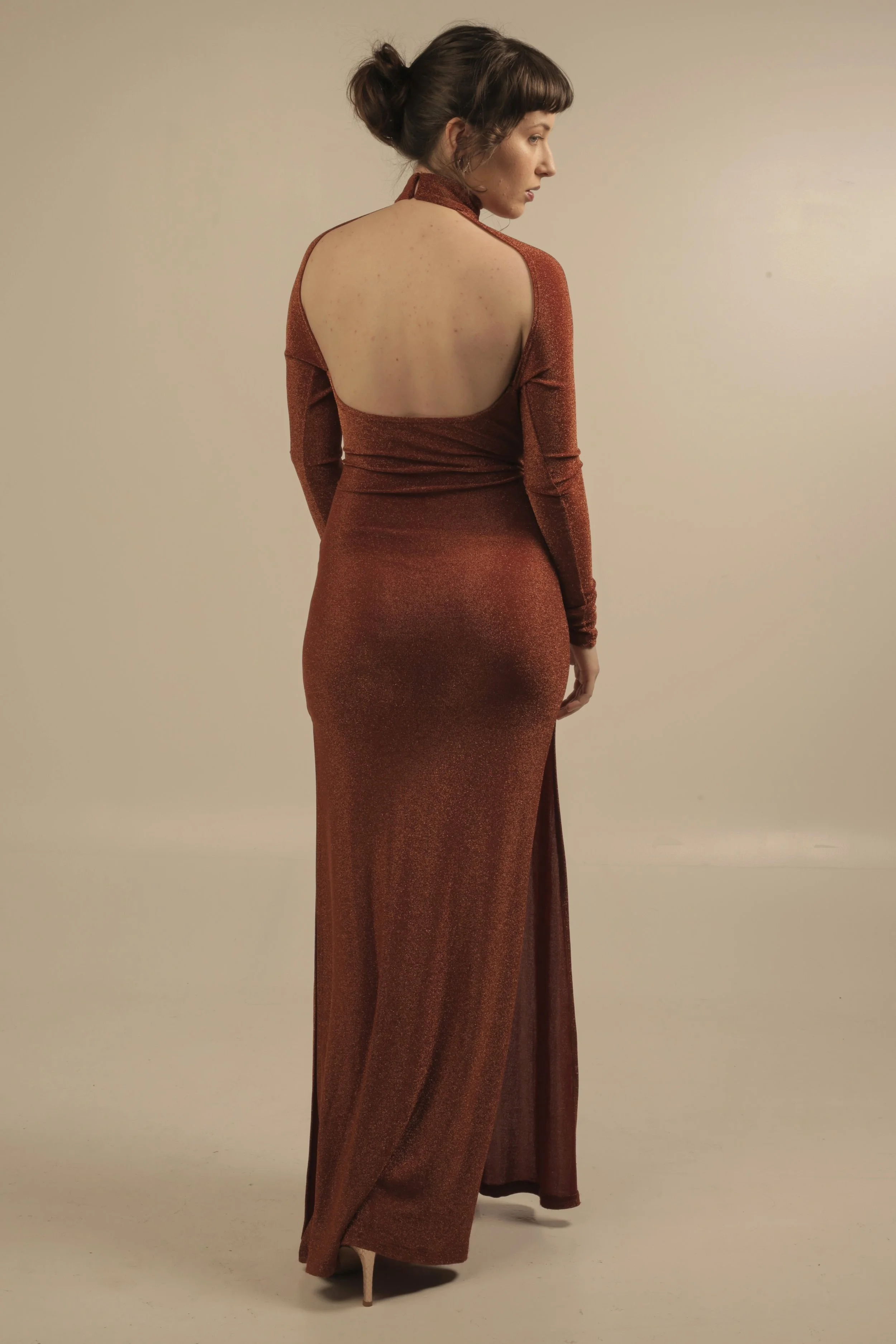 Woman wearing a long, fitted, shimmery, rust-colored gown with an open back, standing against a beige background, looking to her right.