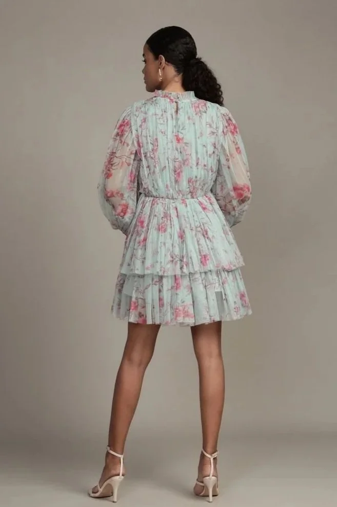 A woman standing with her back to the camera, wearing a flowy, floral dress with long sleeves and high heels, against a plain background.