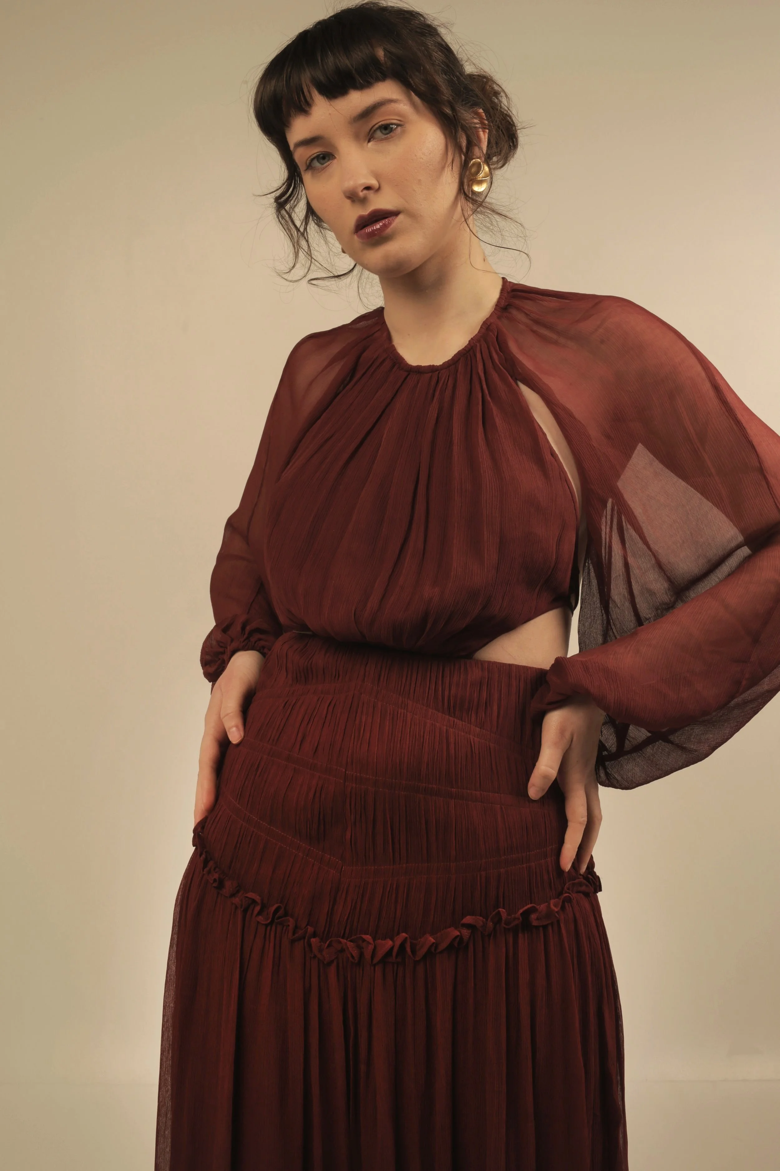 A woman with short dark hair wearing a sheer, pleated, long-sleeve, burgundy dress with cutouts at the waist and a wide skirt, standing against a plain beige background.