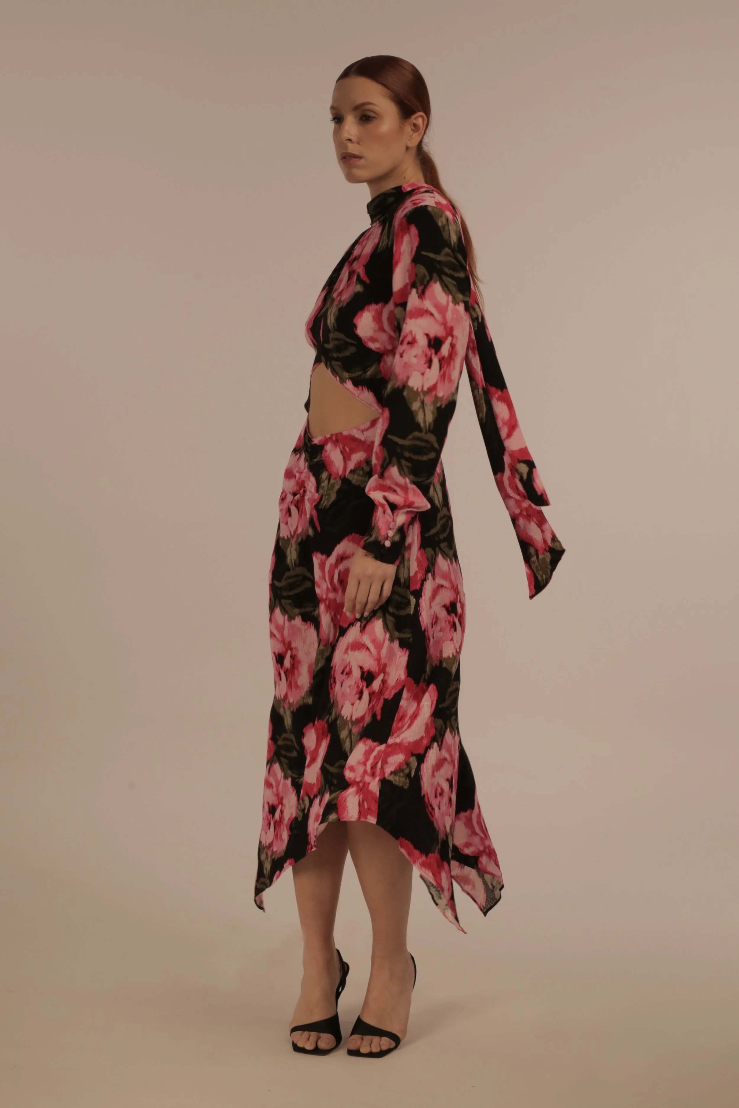 A woman with red hair in a floral dress with cutouts, standing against a plain background, wearing black high heels.