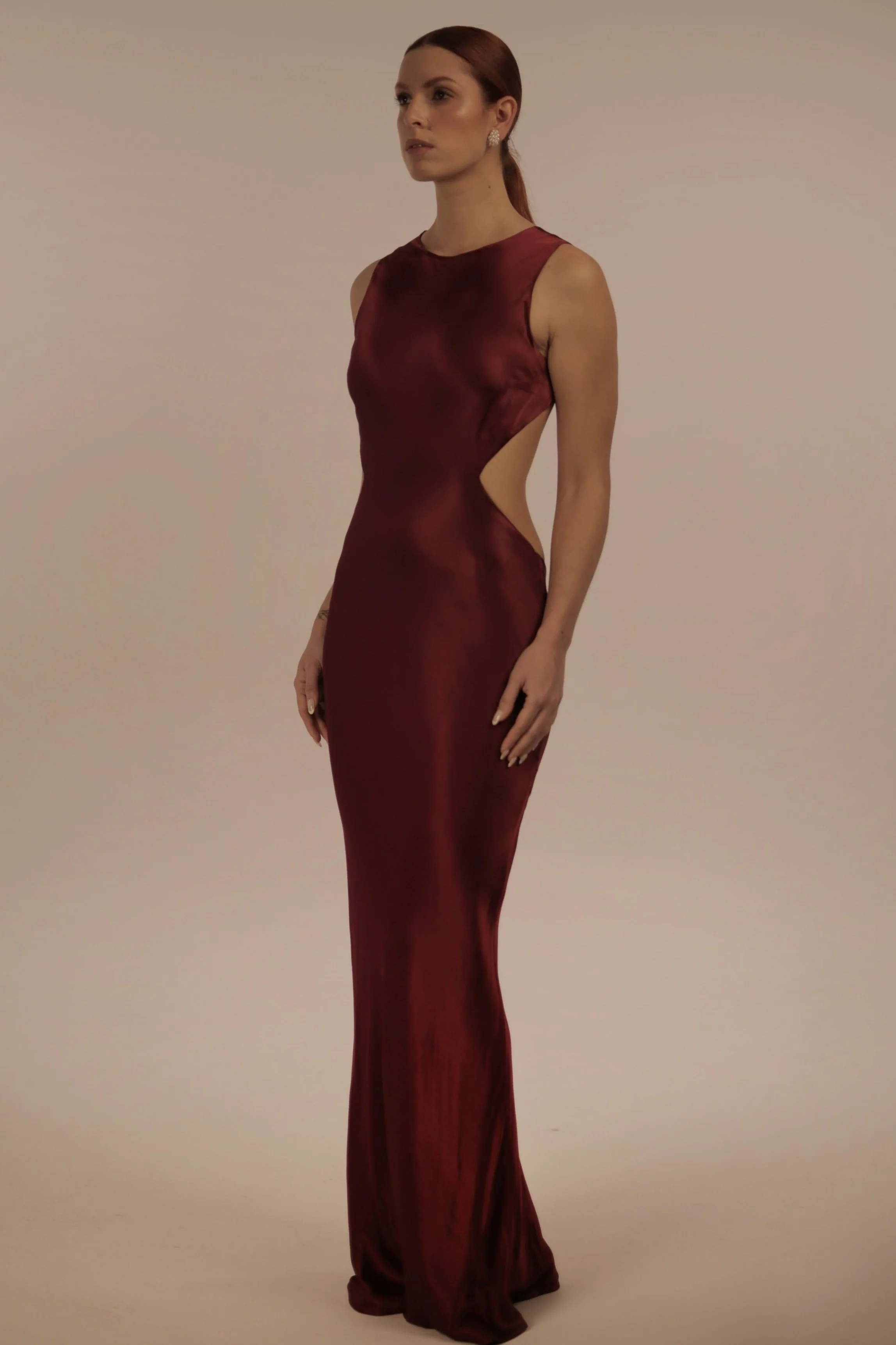 Woman in a long, satin, burgundy gown with side cutouts, standing against a plain background.