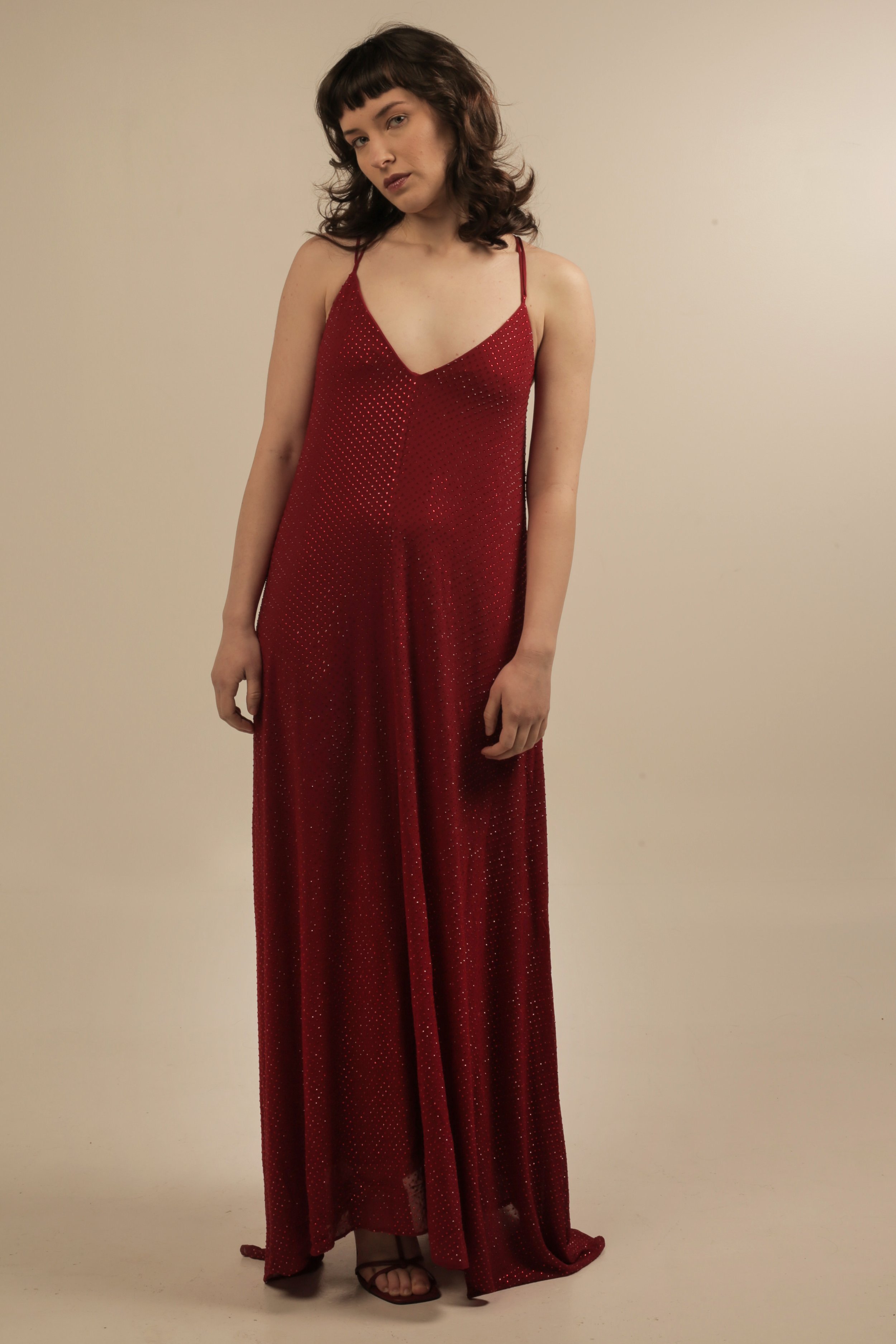 A woman with brown hair and bangs wearing a red, sparkly, sleeveless, floor-length dress with spaghetti straps, standing against a plain beige background.