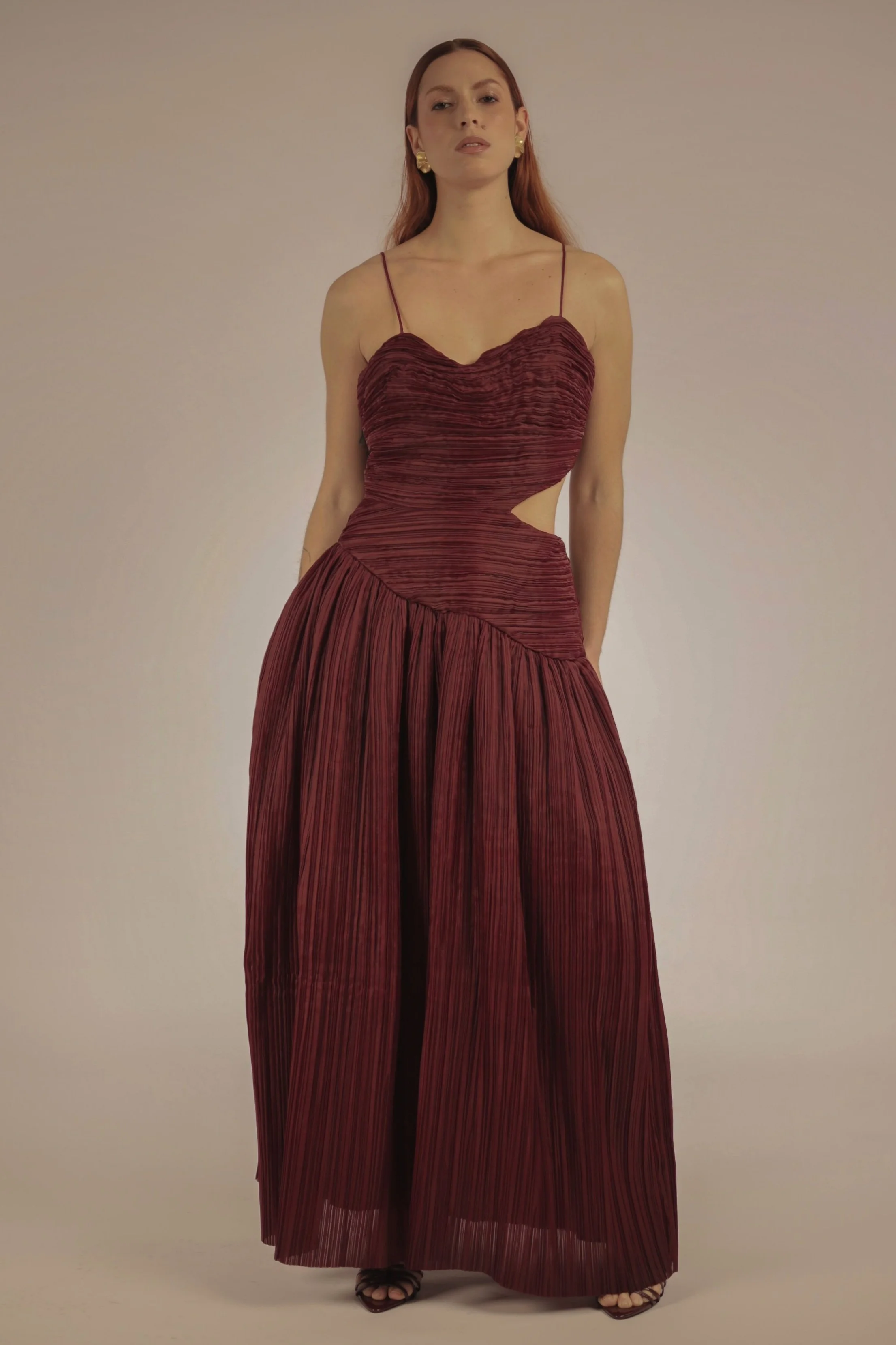 A woman with red hair wearing a deep red pleated dress with cutouts on the sides, standing against a plain background.