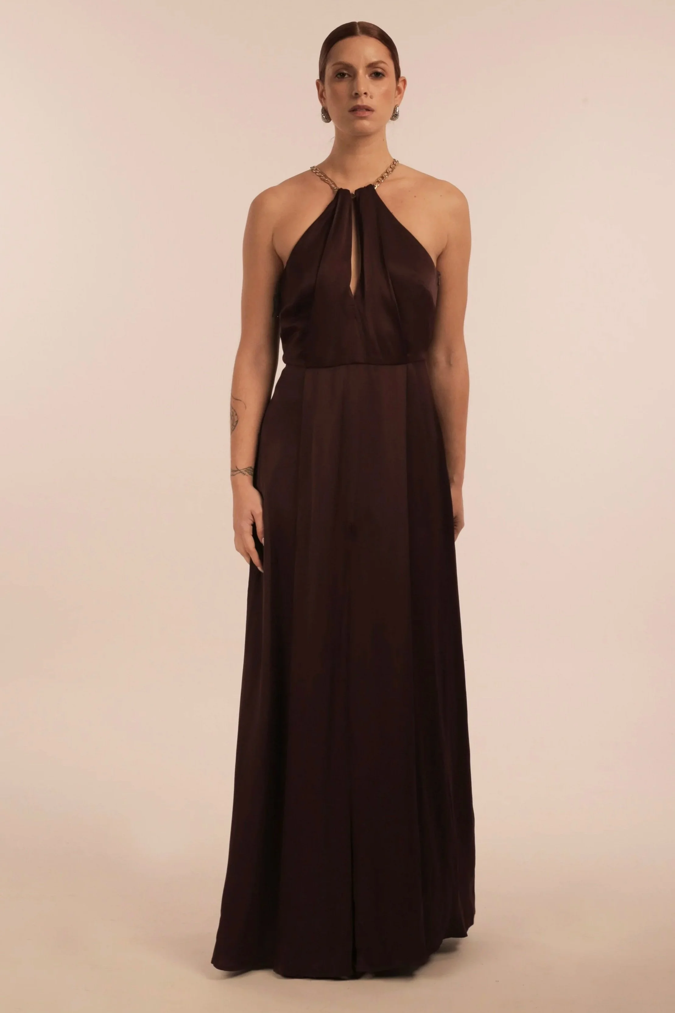A woman wearing a long, dark brown halter gown with a keyhole neckline, standing against a plain, light-colored background.