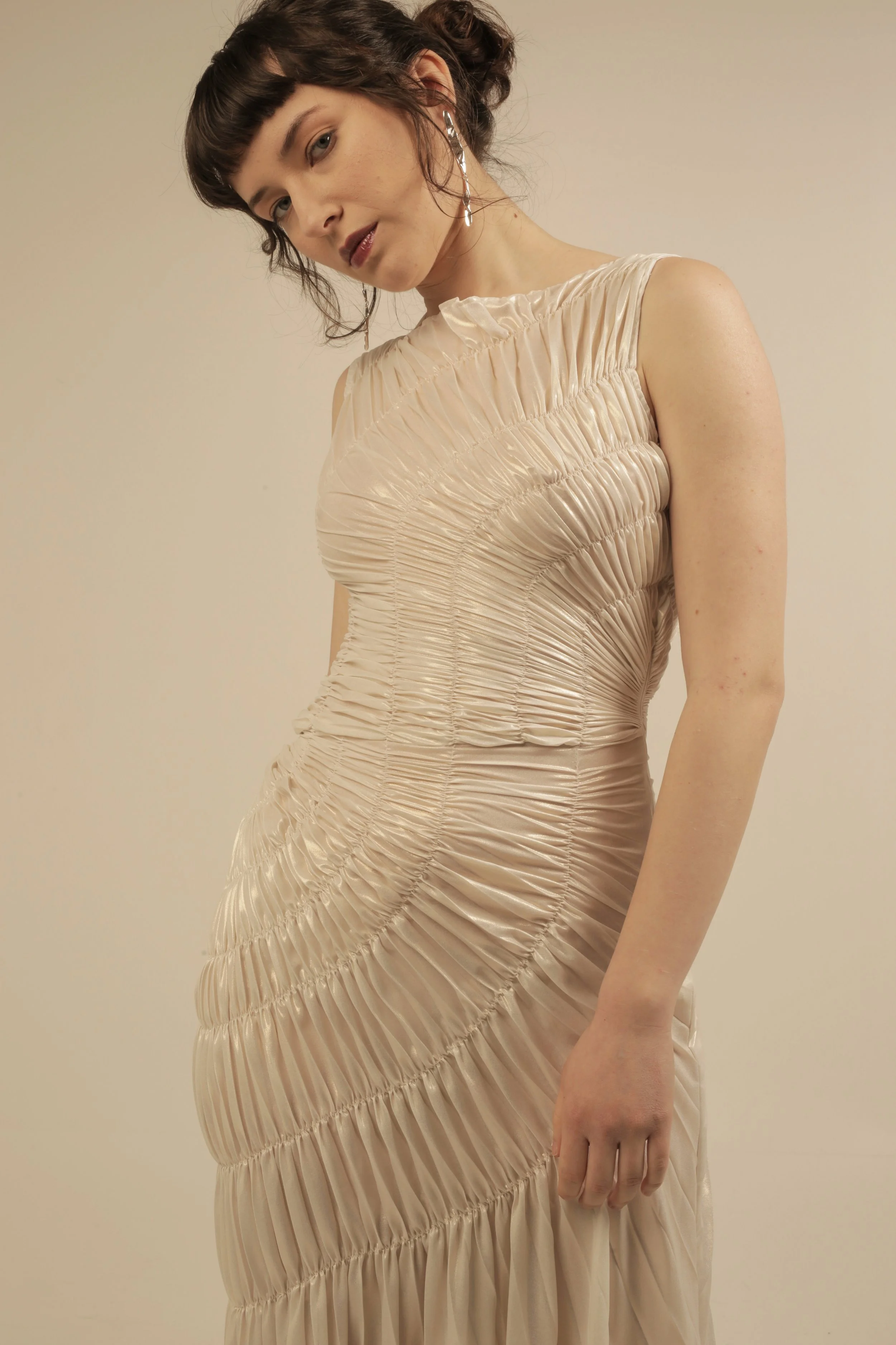 Young woman with dark hair and bangs wearing a light-colored, ruched dress and long earrings, standing against a beige background.
