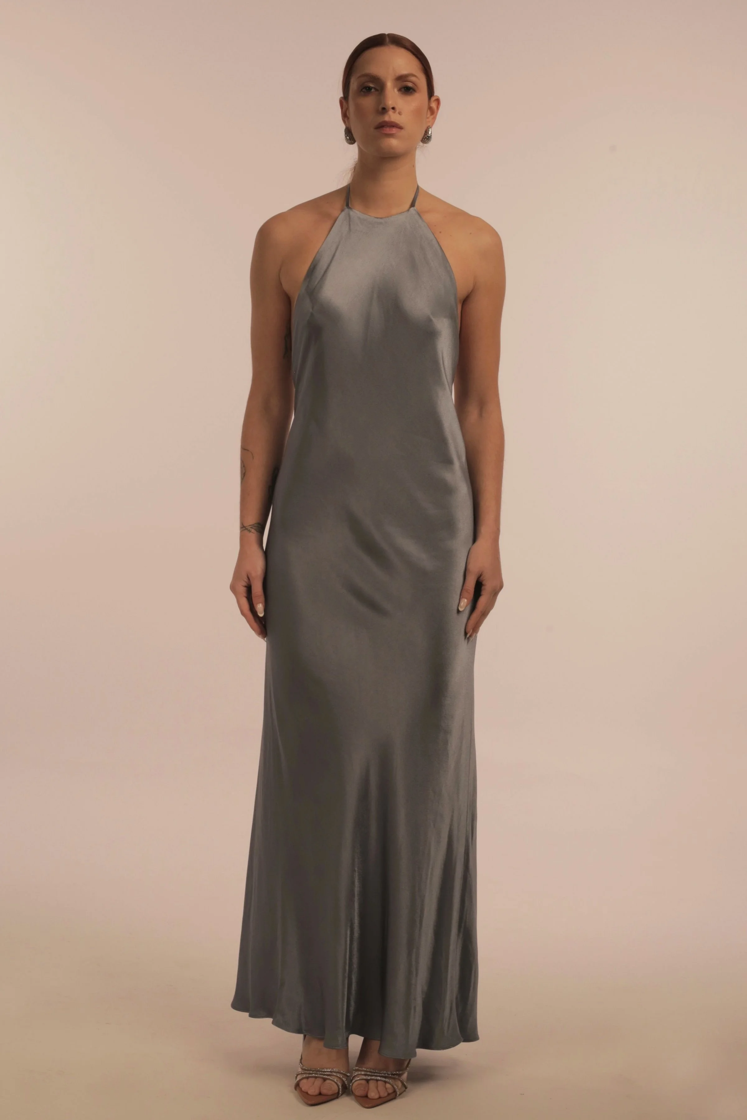 A woman with medium-length dark hair styled back, wearing a long, silver satin halter evening gown and high-heeled sandals, standing against a plain beige background.