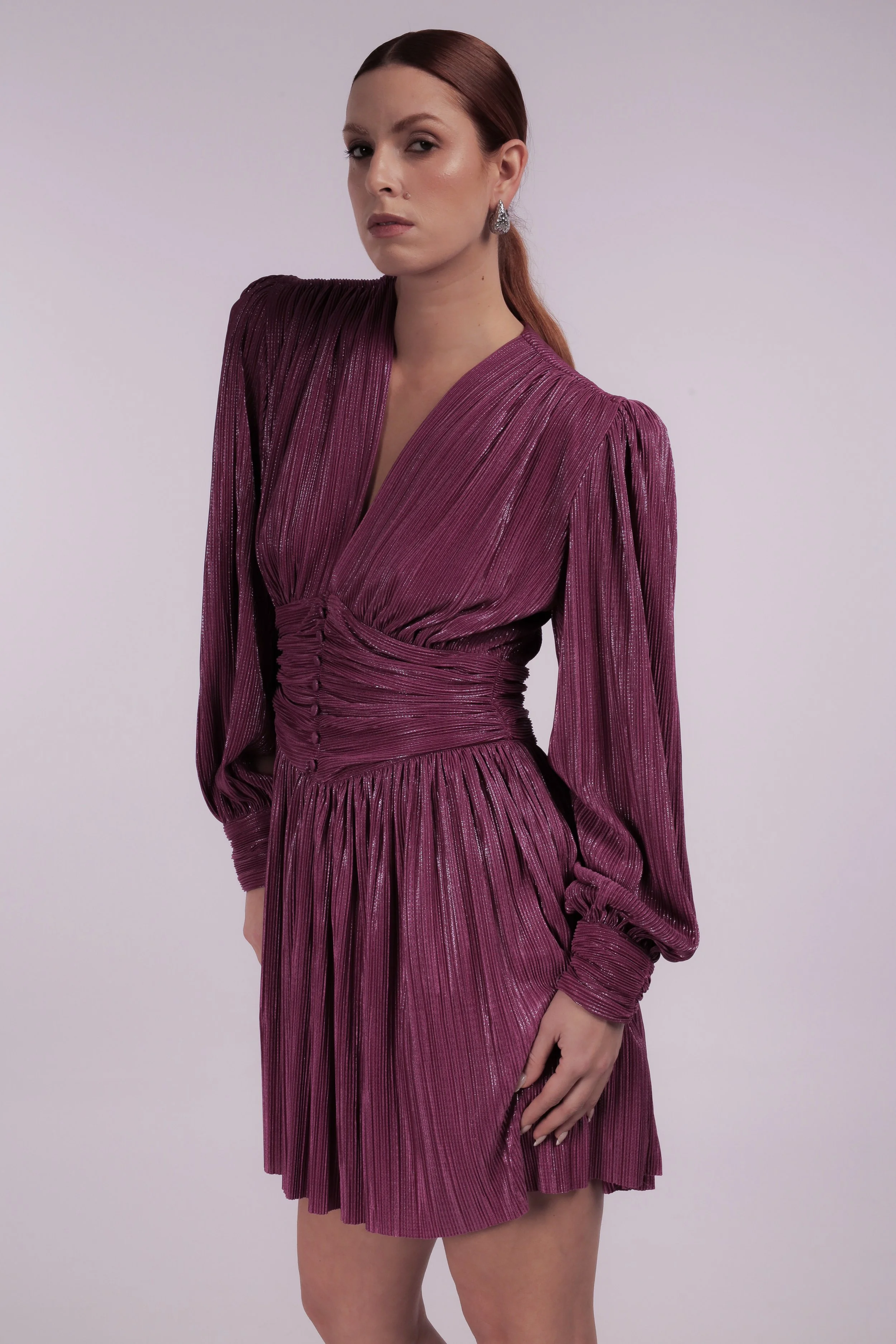 A woman with red hair wearing a shimmery purple pleated dress with puffed sleeves and earrings.