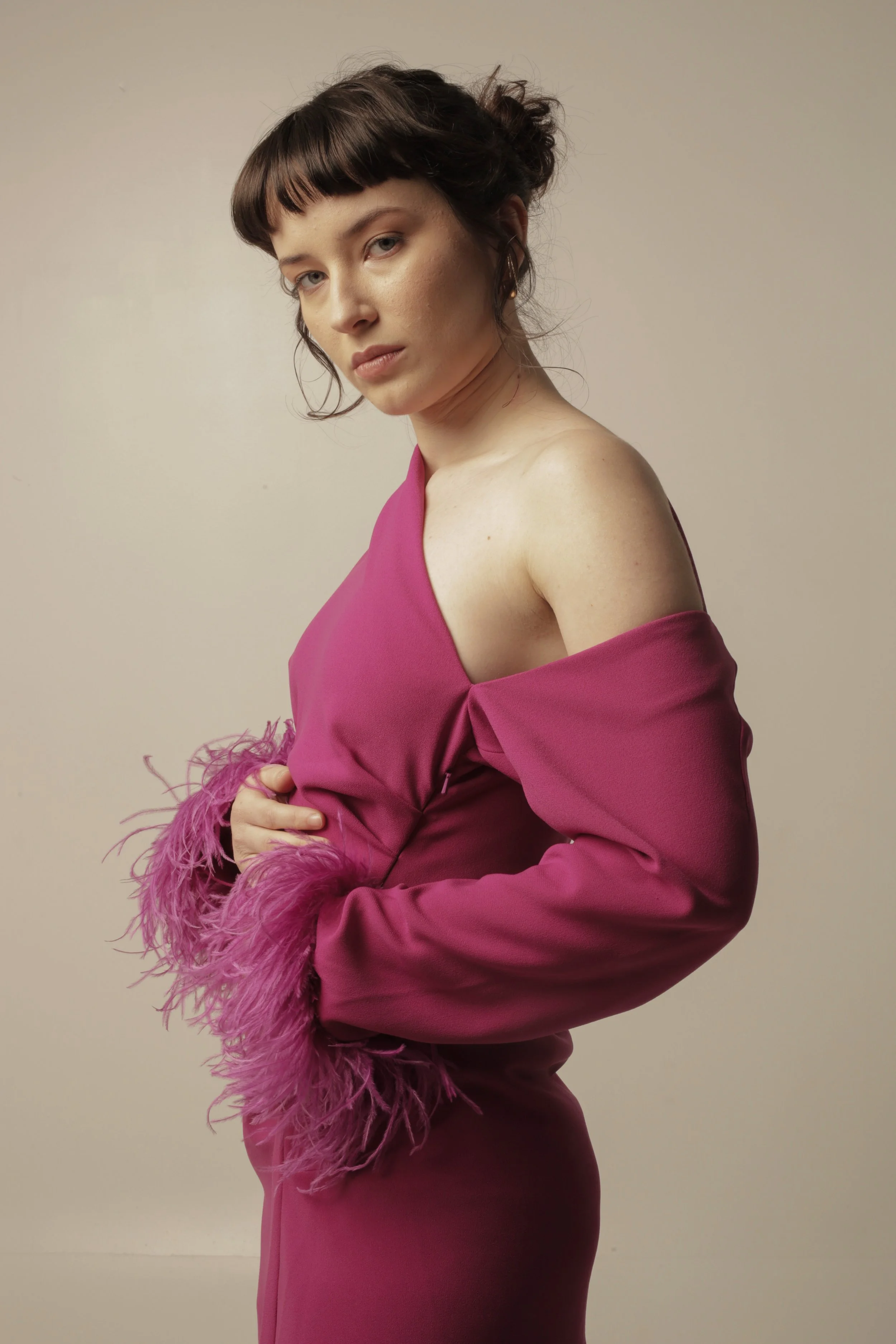 A woman with dark brown hair styled in a bun with bangs, wearing a one-shoulder, fuchsia dress with feathered cuffs, stands against a plain beige background.