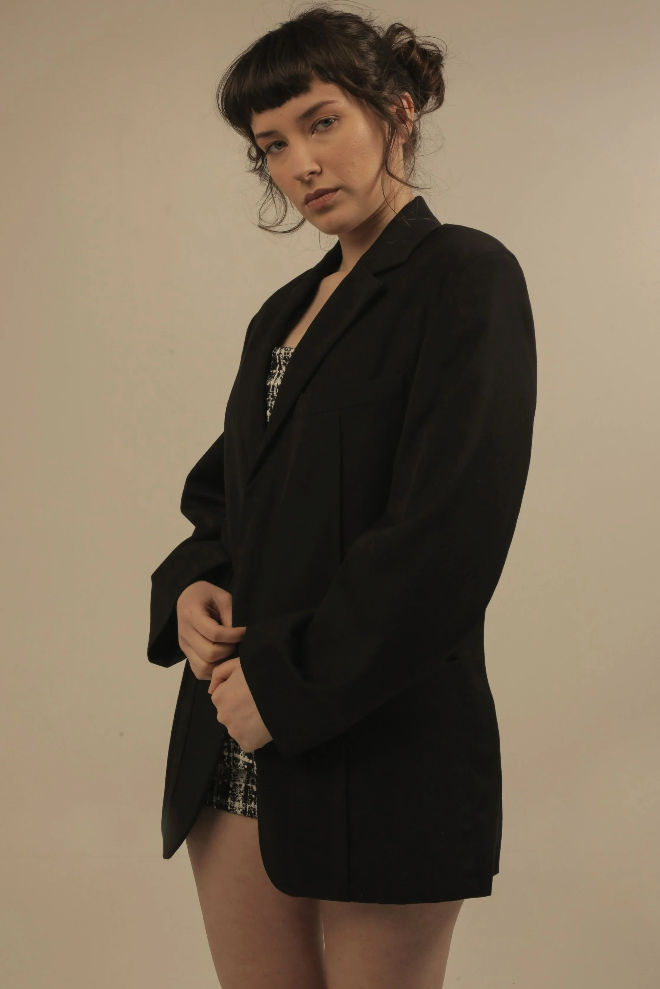 A woman with dark hair styled in a messy bun and bangs, wearing a black blazer over a patterned dress, standing against a plain beige wall.