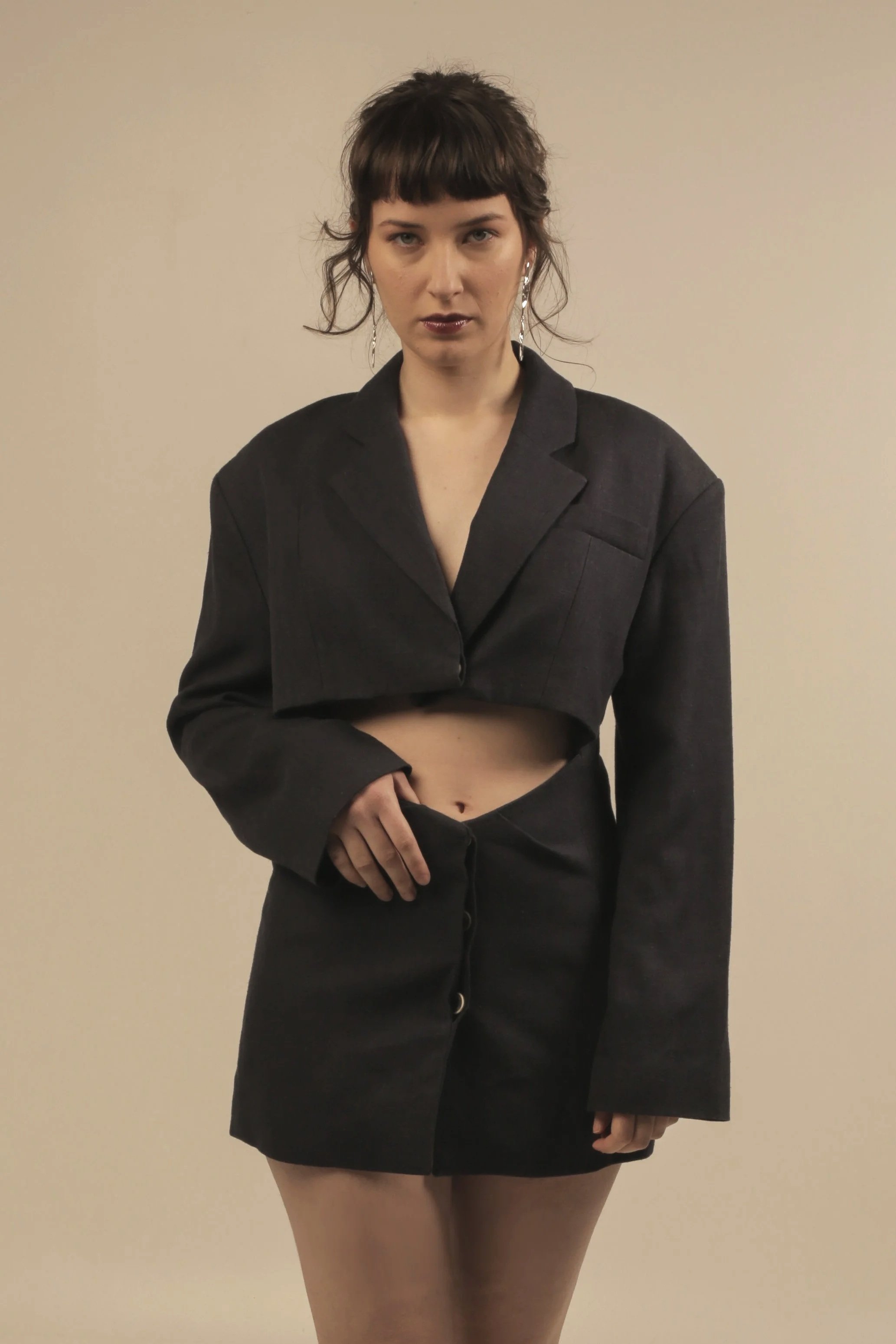 A woman with dark hair, wearing a black blazer and matching skirt, standing against a plain wall.