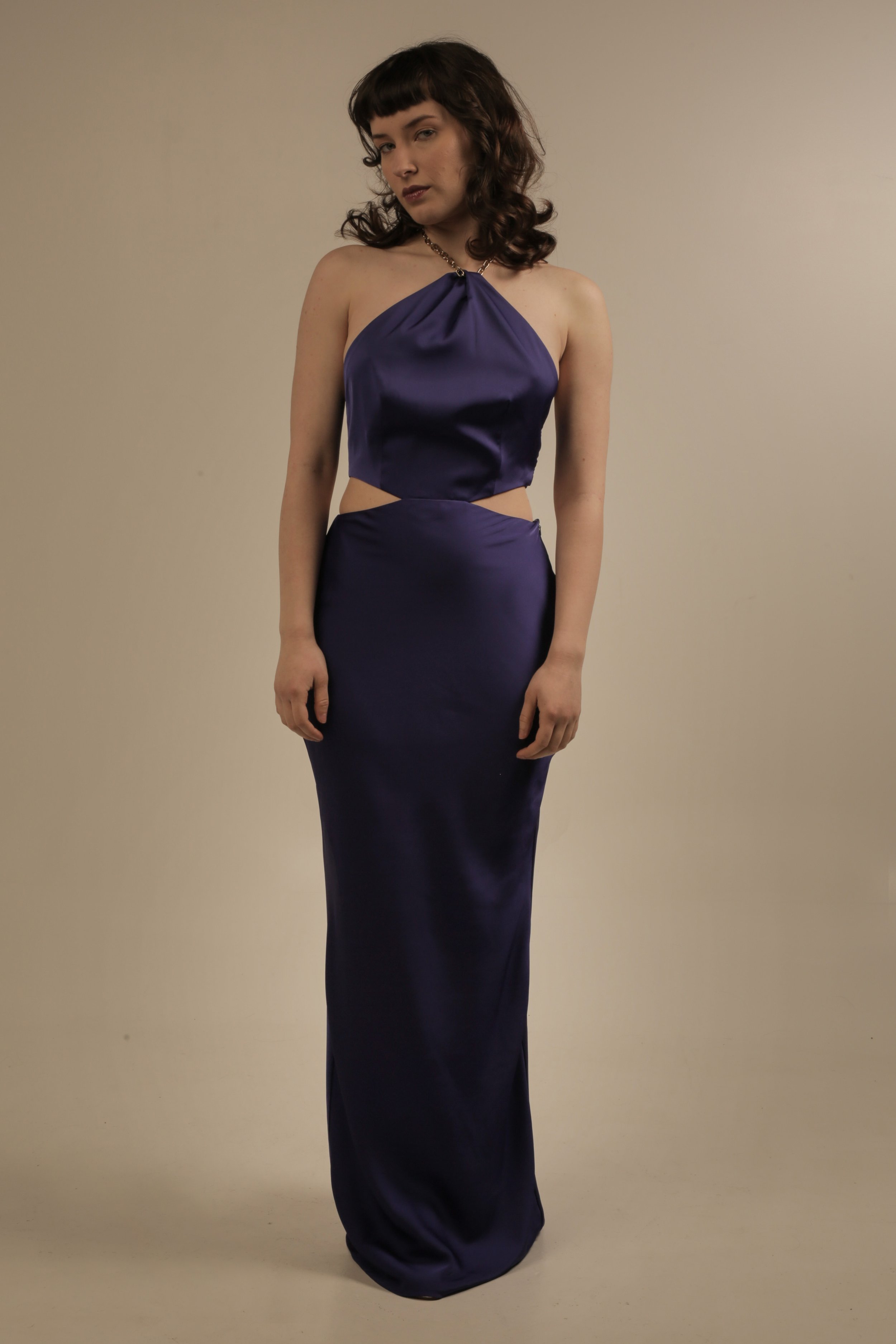 A woman with dark wavy hair wearing a dark purple satin gown with cutouts at the sides and a halter neckline, standing against a beige background.
