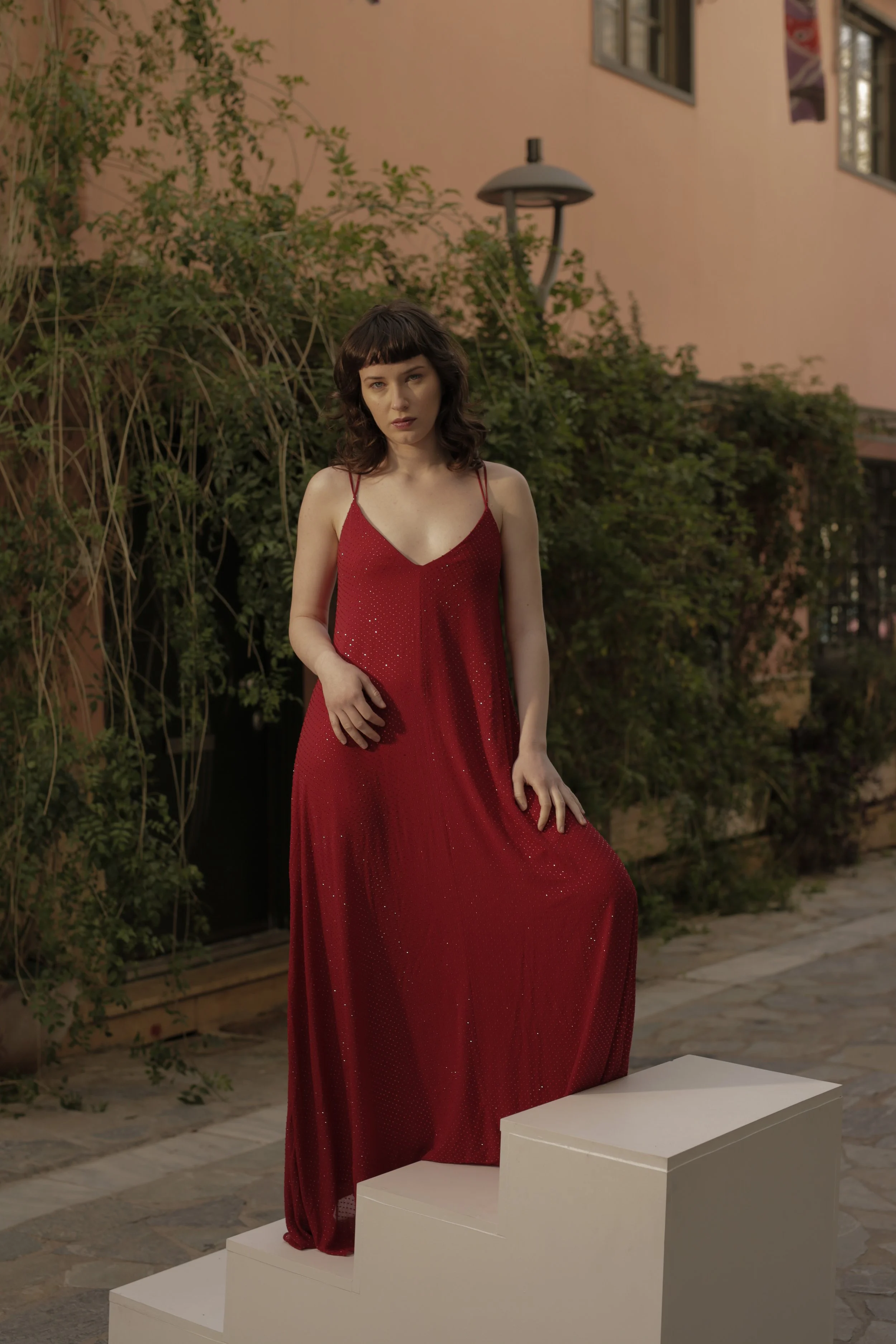A woman in a red dress standing on a three-step white pedestal outdoors, with a peach-colored building and green bushes in the background.