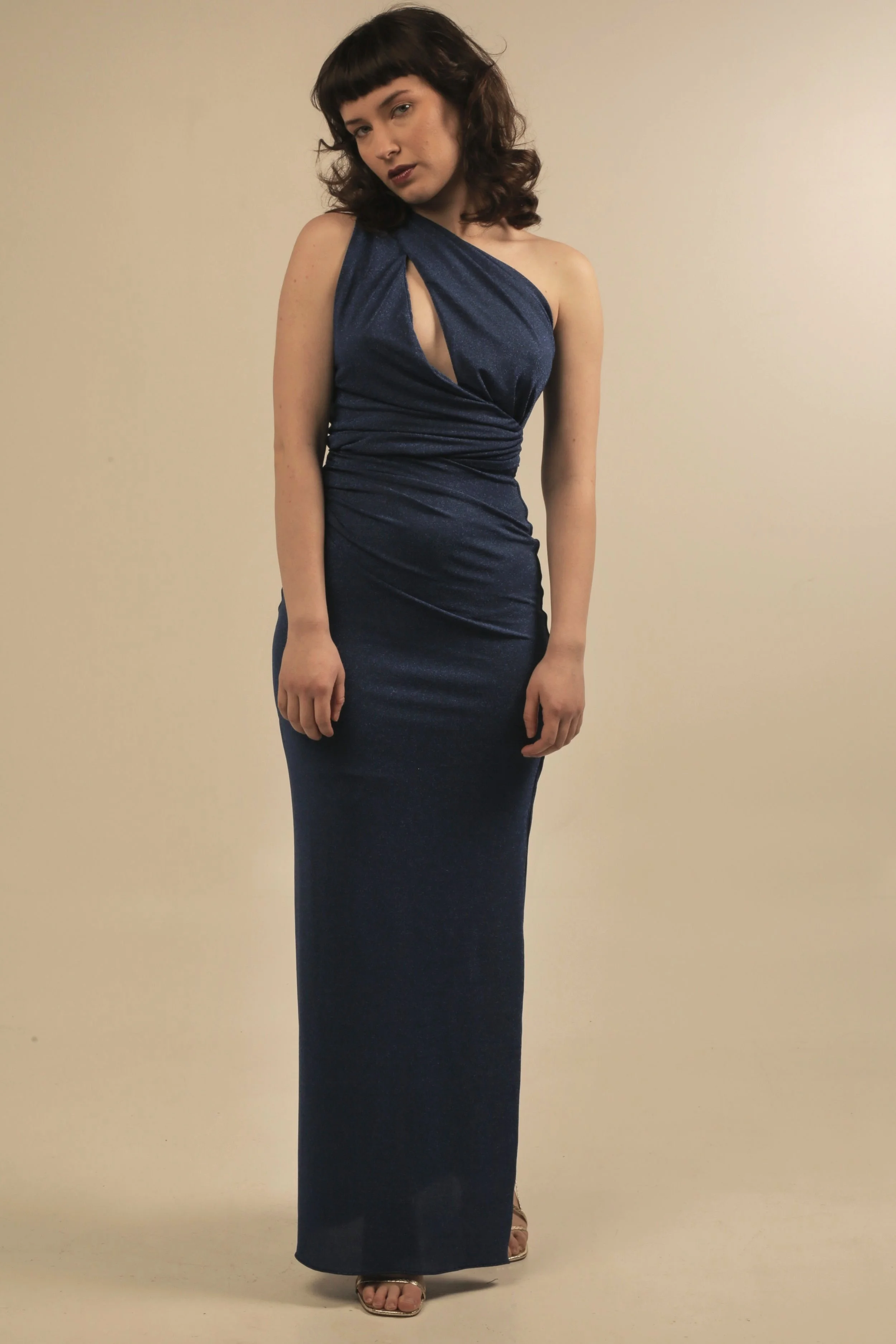 Woman in a dark blue, floor-length, one-shoulder dress with a cutout detail, standing against a plain light-colored background.