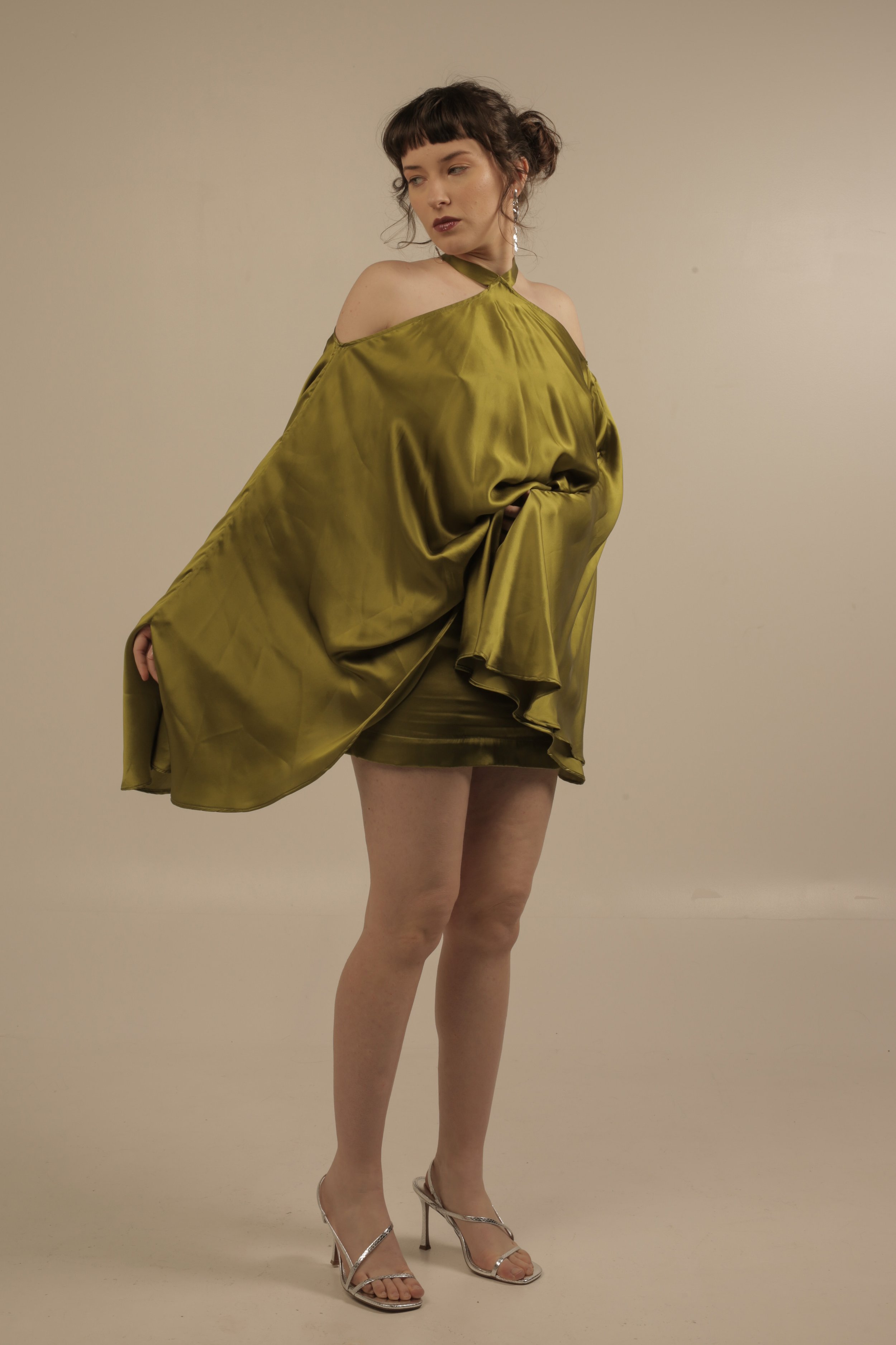 A woman standing against a plain background, wearing a shiny olive green off-shoulder dress, silver high heels, and dangling earrings.