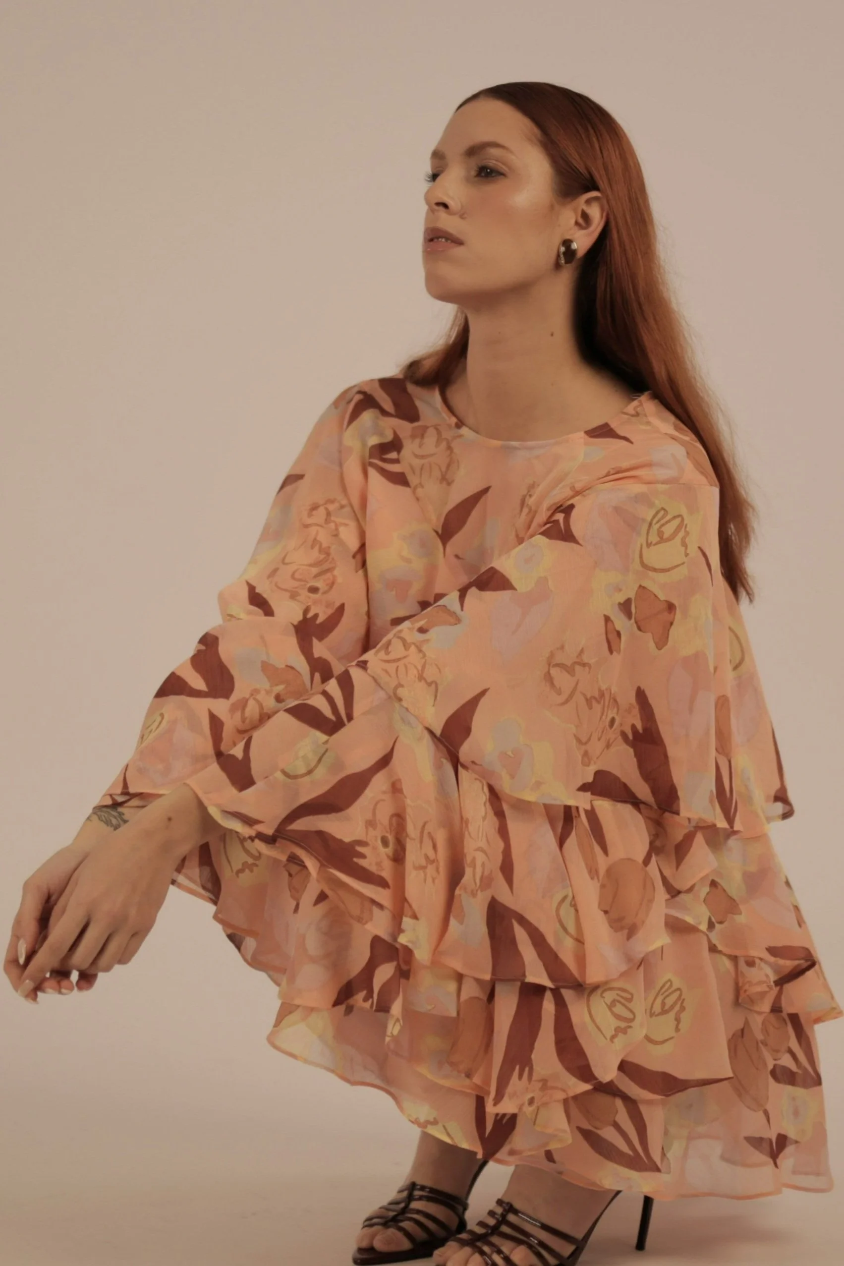 A woman with long red hair wearing a peach-colored floral dress and black strappy high heels, sitting with her arms crossed against a plain light-colored background.